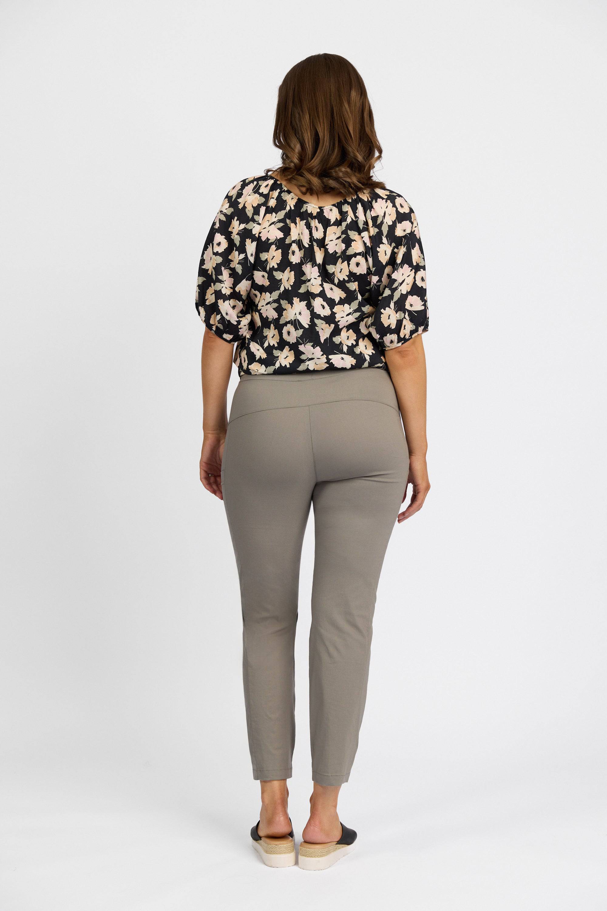 A person with shoulder-length brown hair faces away, wearing a black floral top, Vassalli Paris Cargo Capri pants in Latte, and black slip-on shoes. The background is plain white.