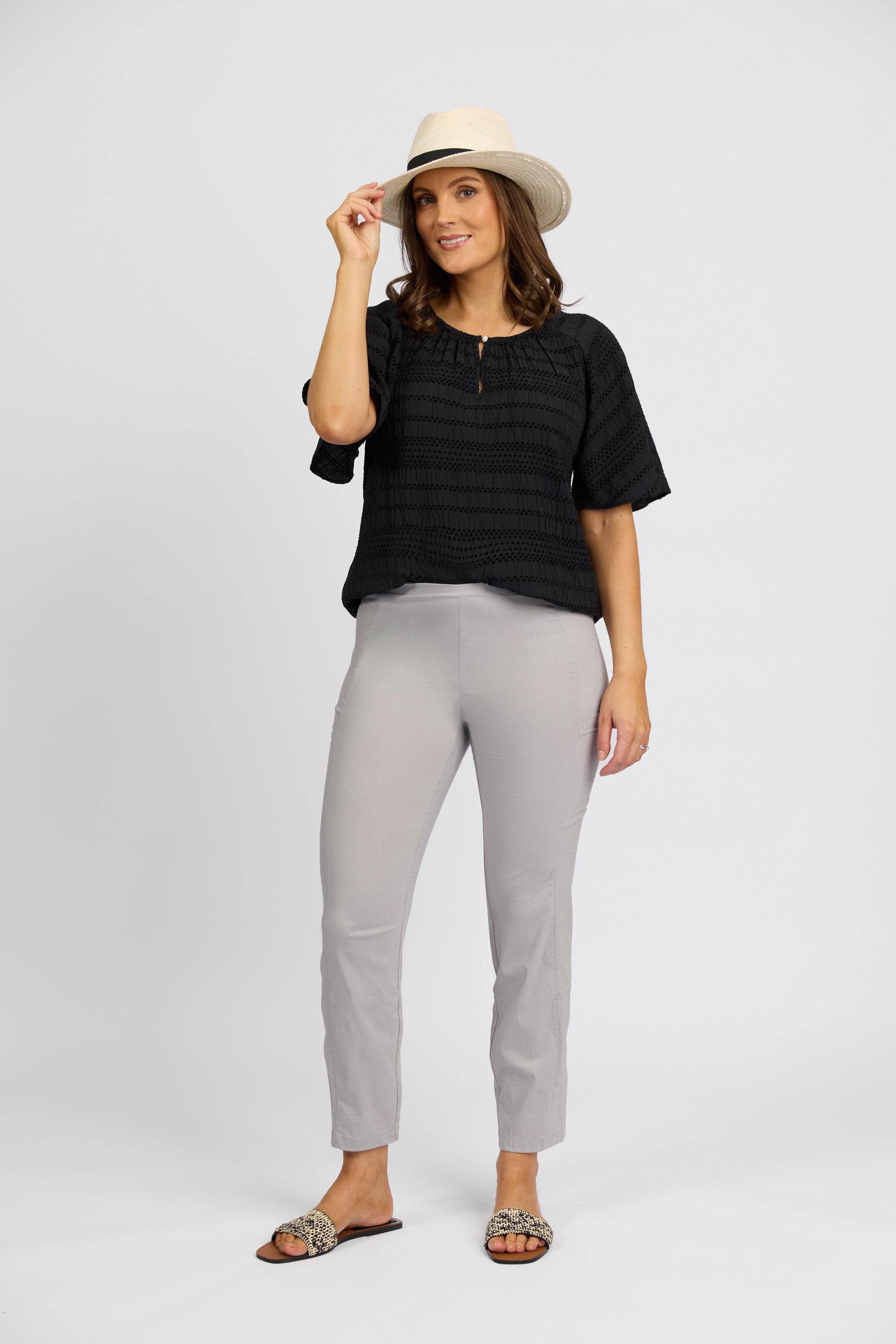 A woman stands against a plain background, smiling and holding her hat, dressed in a black short-sleeve top, Vassalli Paris Cargo Capri - Silver, sandals, and a light-colored hat.