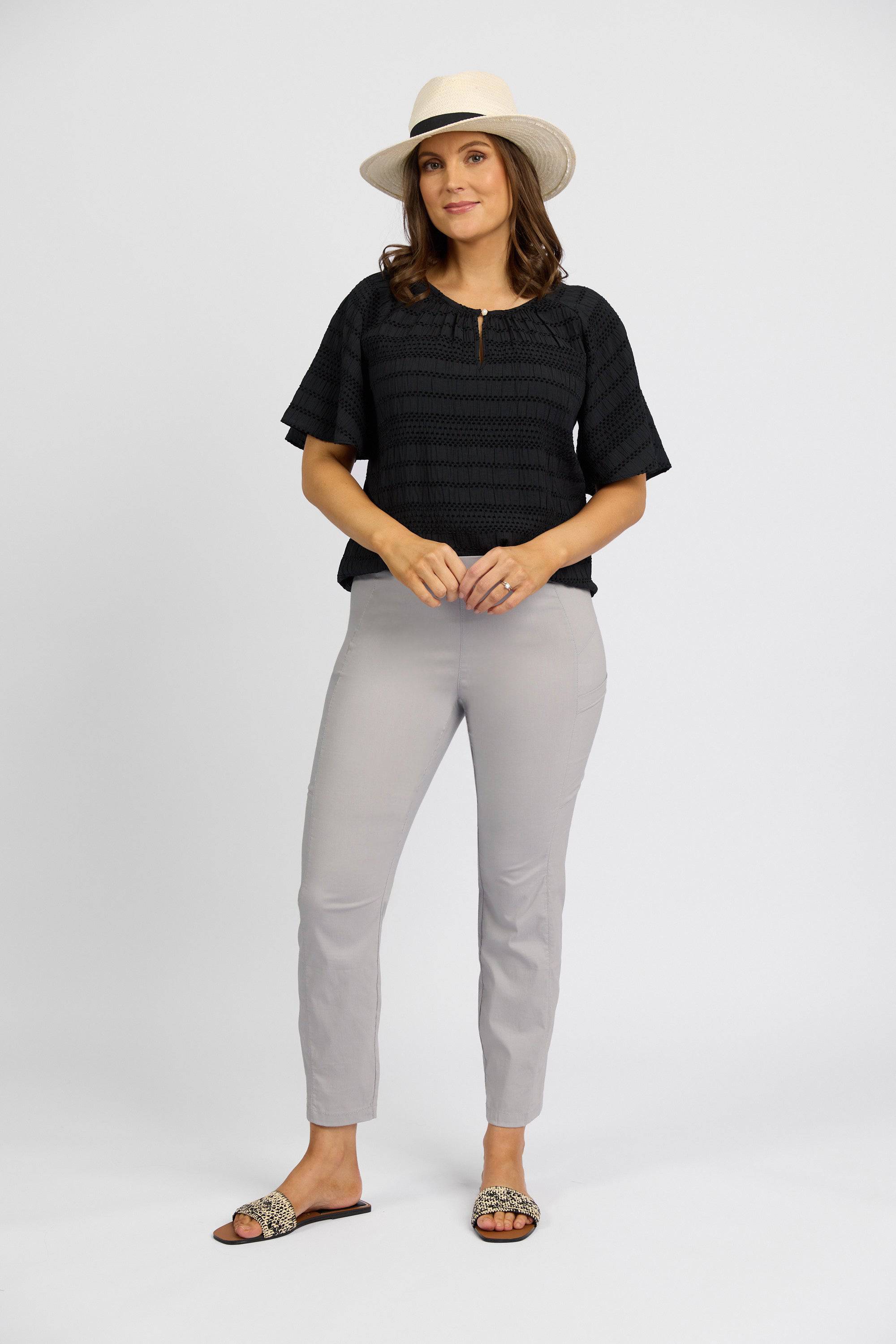 A woman in a straw hat, black short-sleeve top, Vassalli Paris Cargo Capri - Silver, and leopard-print sandals stands against a plain white background, facing the camera with a relaxed expression.
