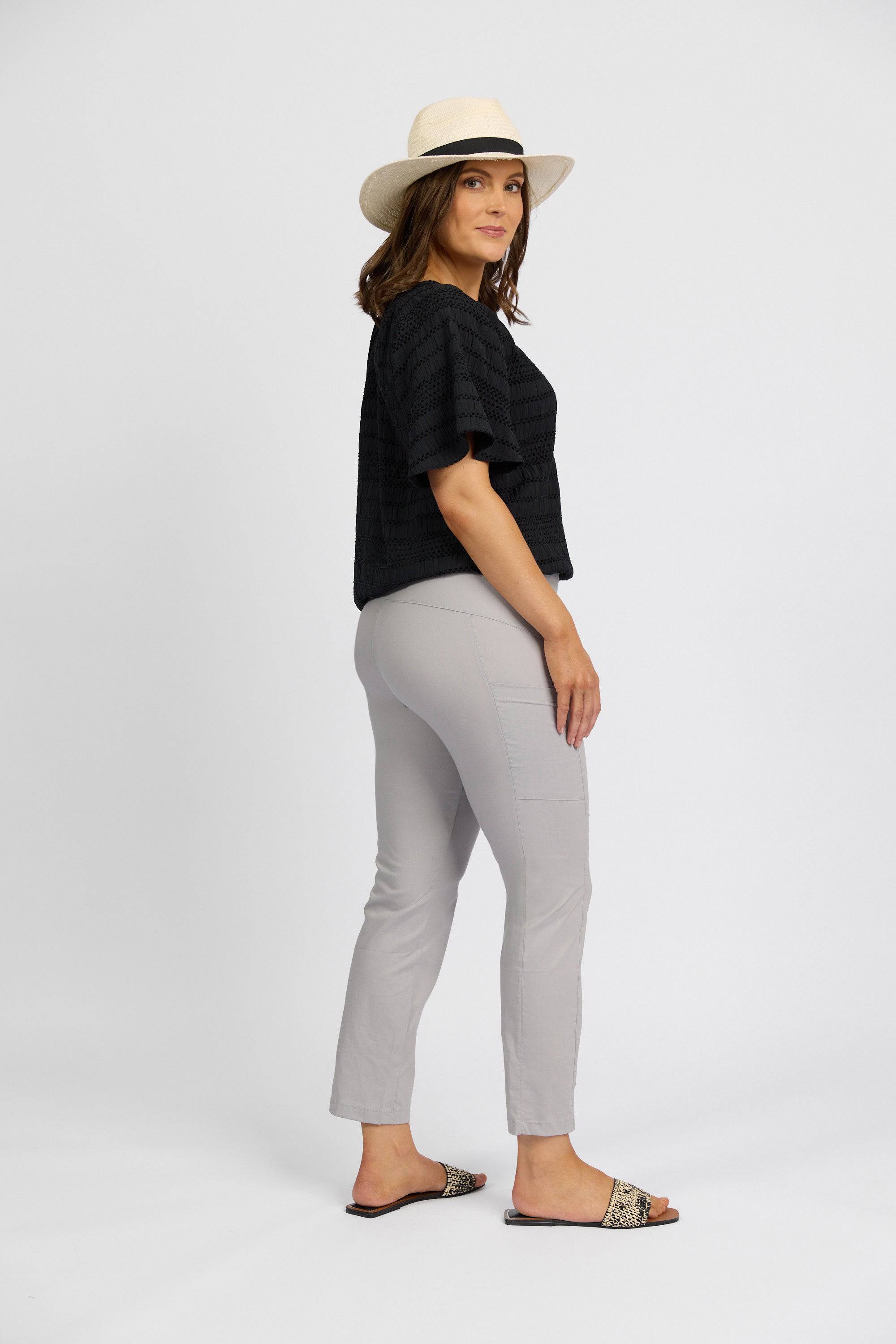 A woman stands sideways facing the camera, wearing a Vassalli black short-sleeve top, Paris Cargo Capri - Silver by Vassalli, a straw hat, and slip-on sandals against a plain white background.