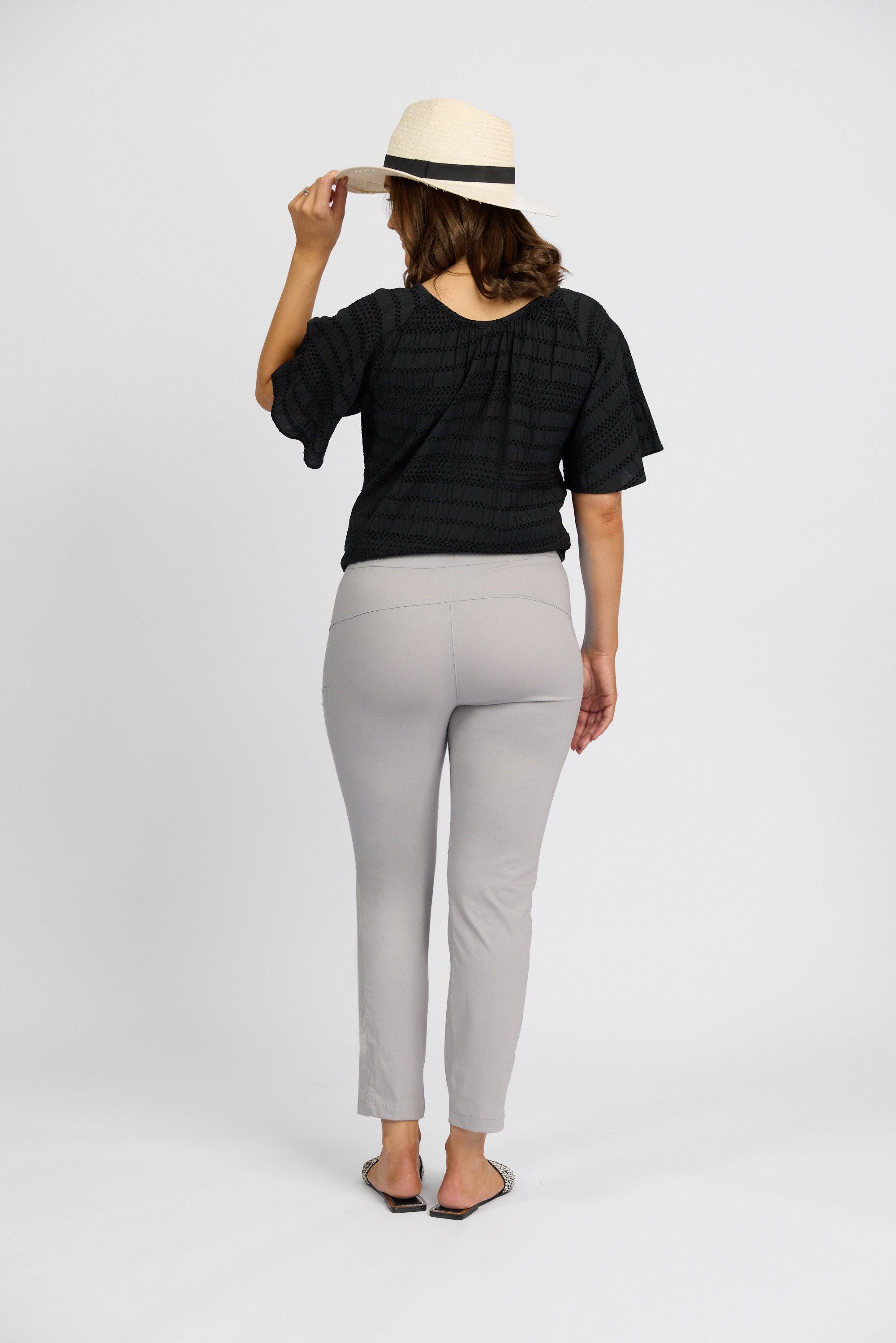 A woman stands facing away, wearing a straw hat, black short-sleeve top, Vassalli Paris Cargo Capri - Silver pants, and flat shoes against a plain white background.