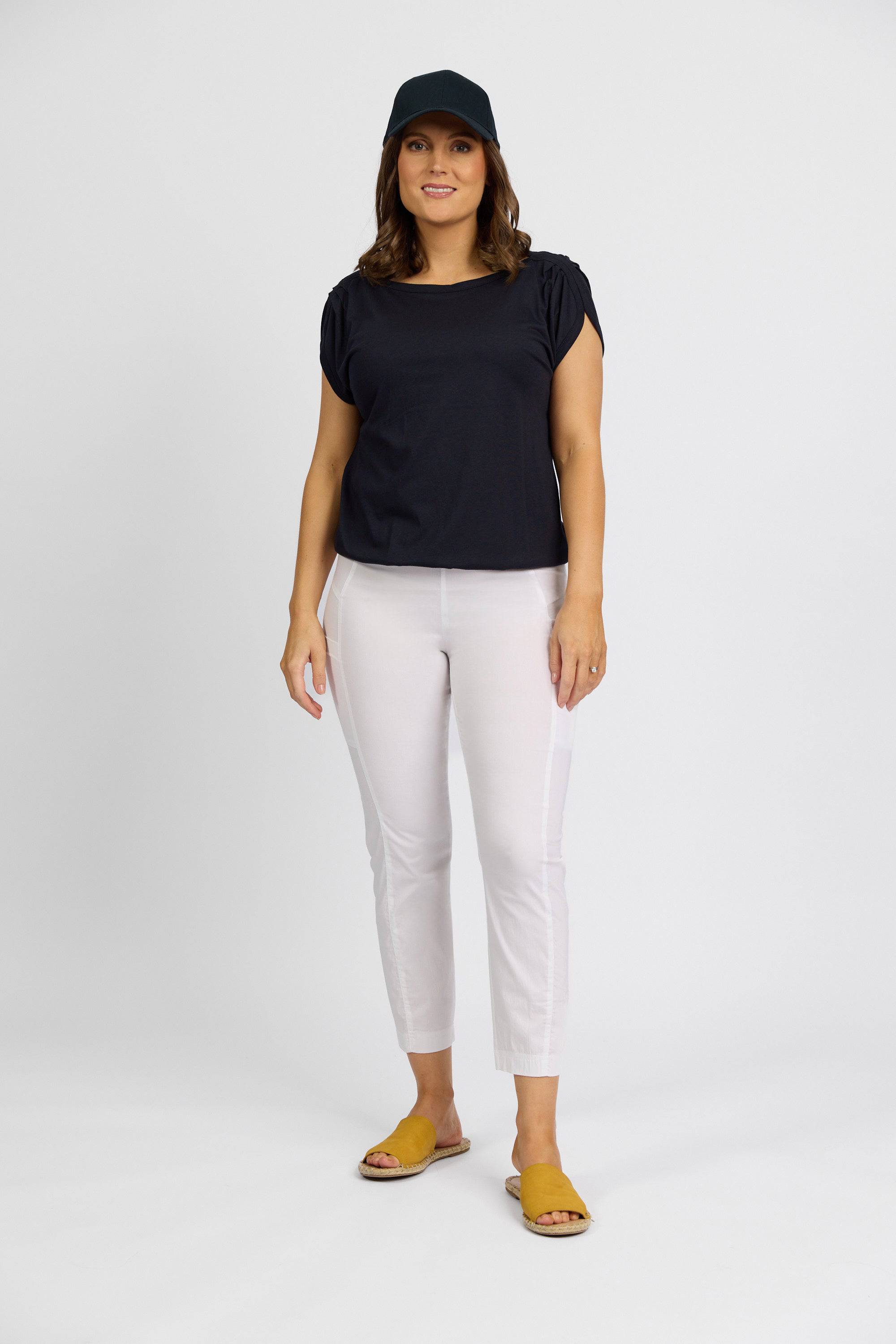 A woman stands against a plain background wearing a black cap, black short-sleeve top, Vassalli Paris Cargo Capri in white, and yellow sandals. She smiles slightly with one hand relaxed by her side.