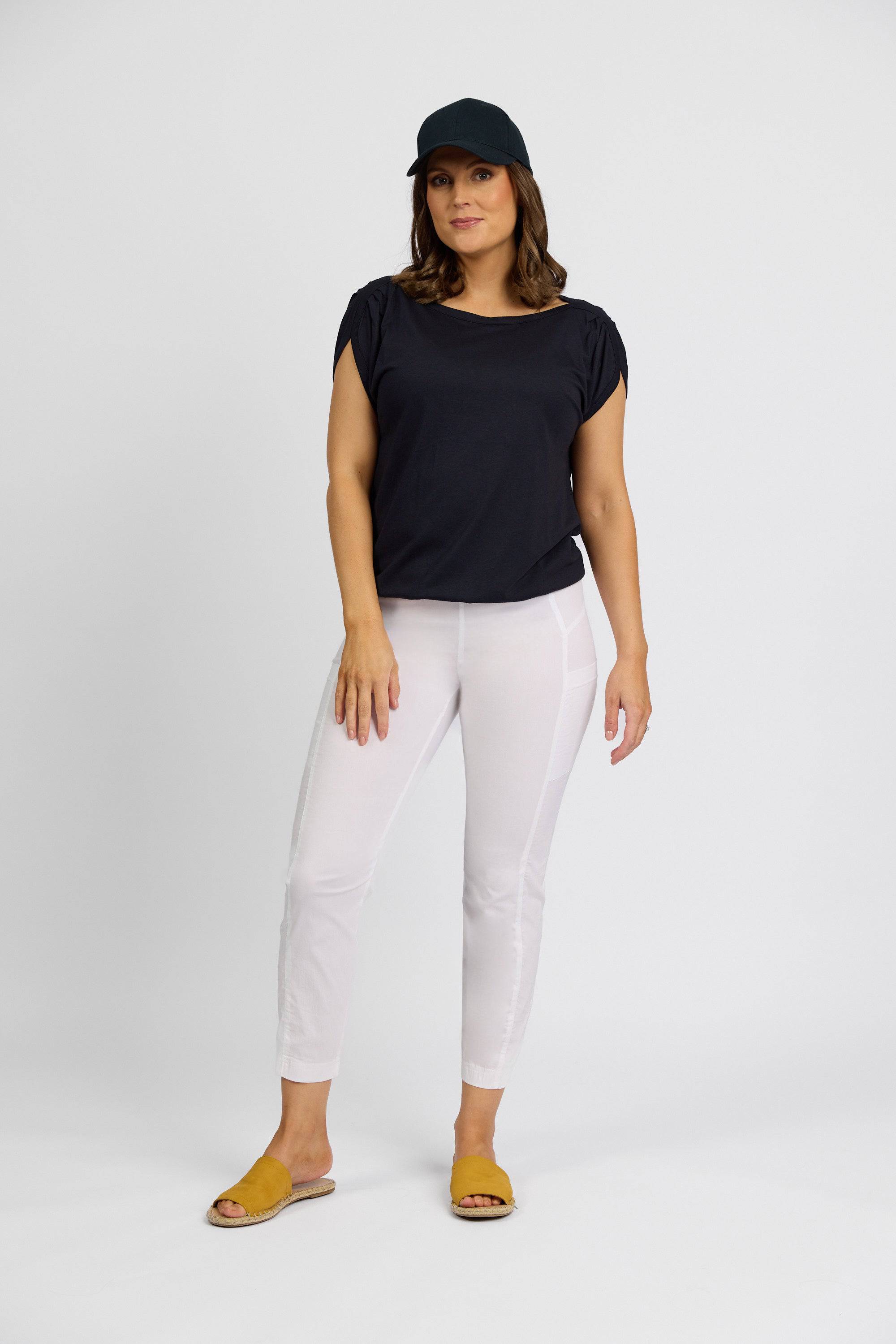 A woman stands against a plain white background wearing a black cap, black short-sleeve top, yellow sandals, and Vassalli Paris Cargo Capri - White. She has one hand relaxed at her side and maintains a neutral expression.