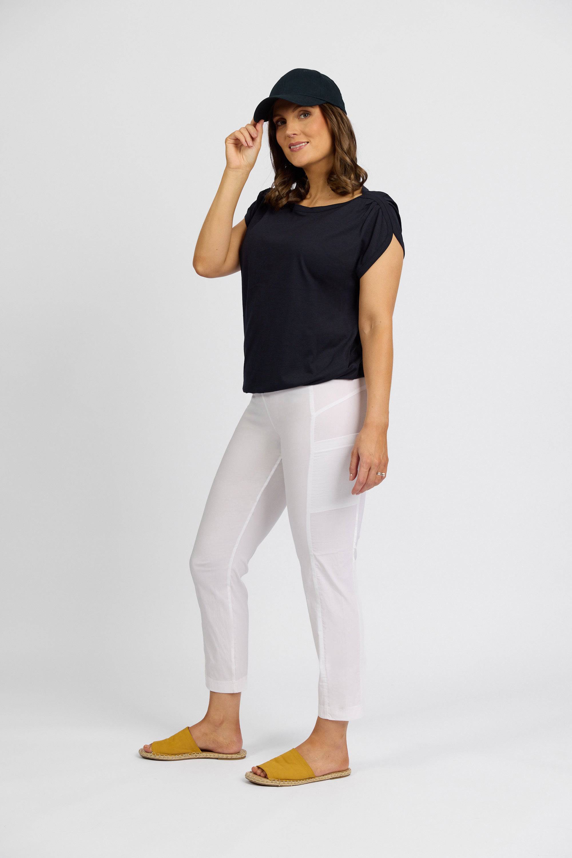 A woman stands against a white background, wearing a black cap and top, Vassalli Paris Cargo Capri in white, and yellow slip-on shoes. She touches her cap’s brim with one hand and smiles slightly.