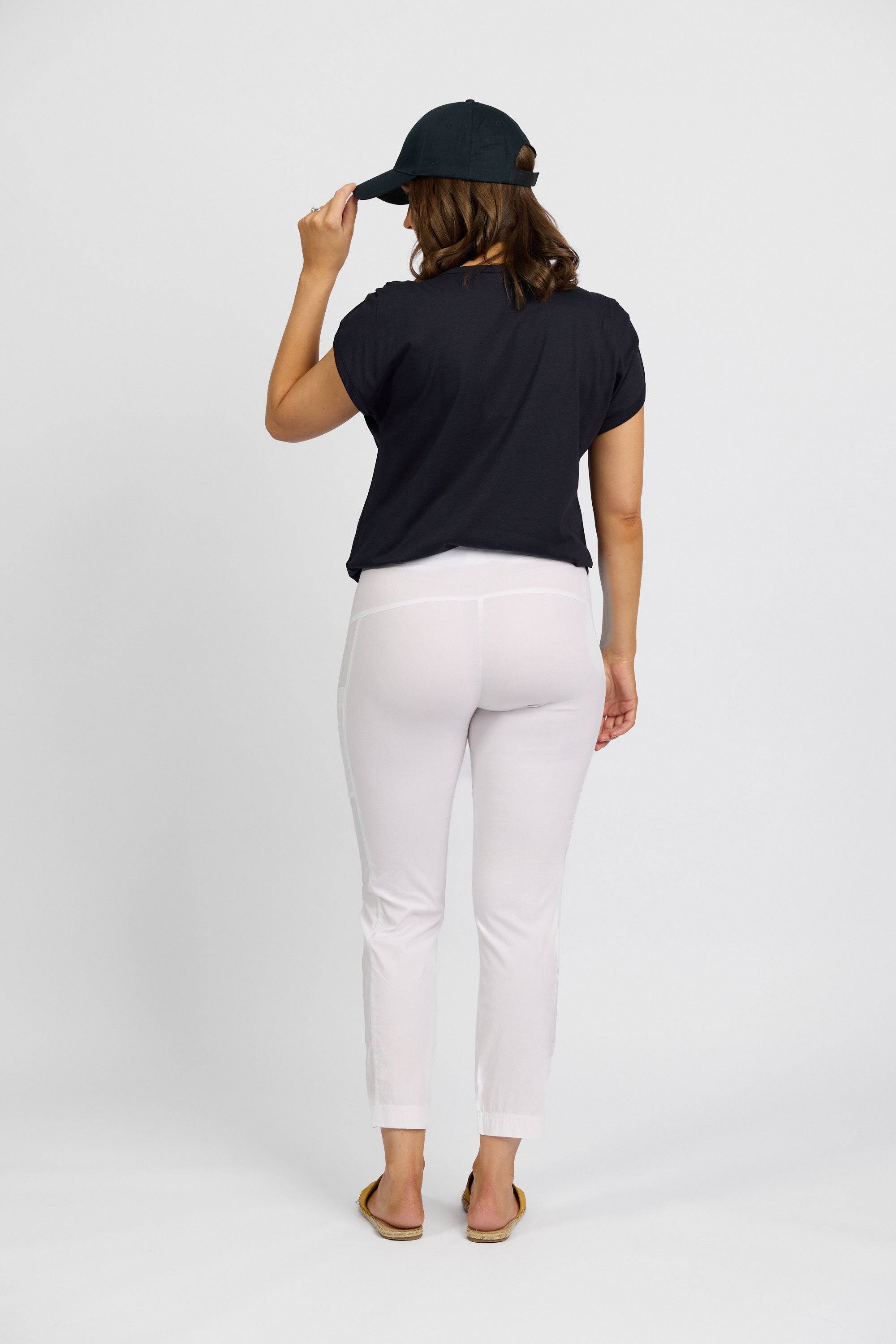 A person stands facing away on a plain white background, wearing a black cap, black T-shirt, Vassalli Paris Cargo Capri - White, and sandals. Their right hand holds the brim of the cap.