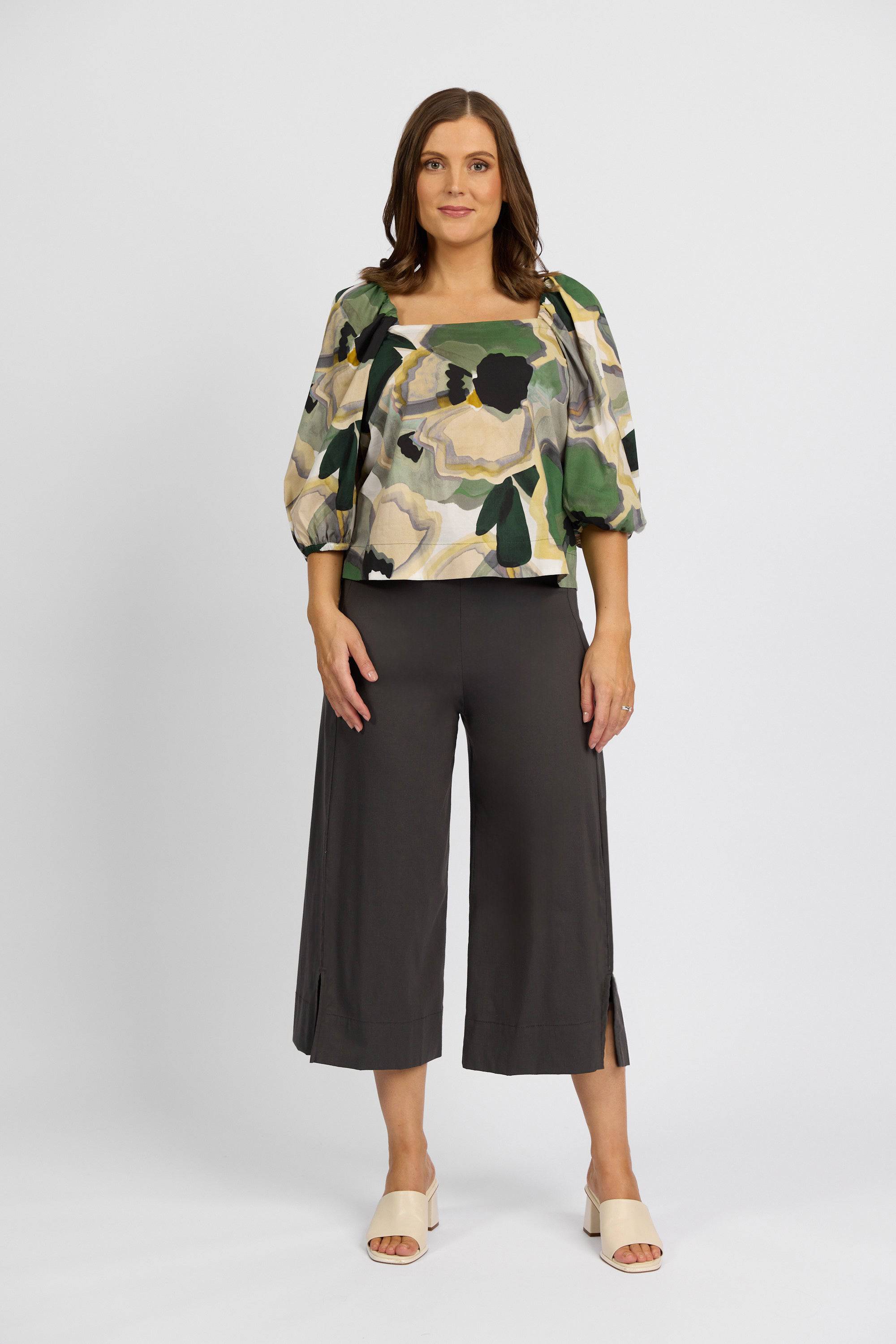 A woman poses against a plain background in a patterned puff-sleeve top and Vassalli's Paris Wide Leg Crop - Beluga pants, made from stretch fabric in dark tones with small side slits, styled with light open-toe sandals.