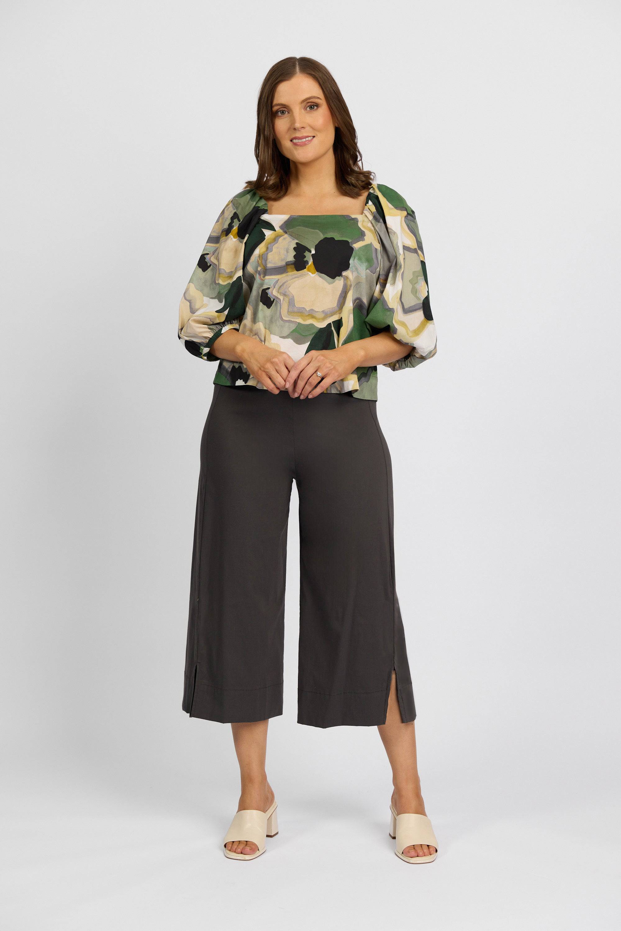 A woman faces forward in a patterned puff-sleeve top, Vassalli Paris Wide Leg Crop - Beluga pants with side slits, and light open-toe sandals, set against a plain white background.
