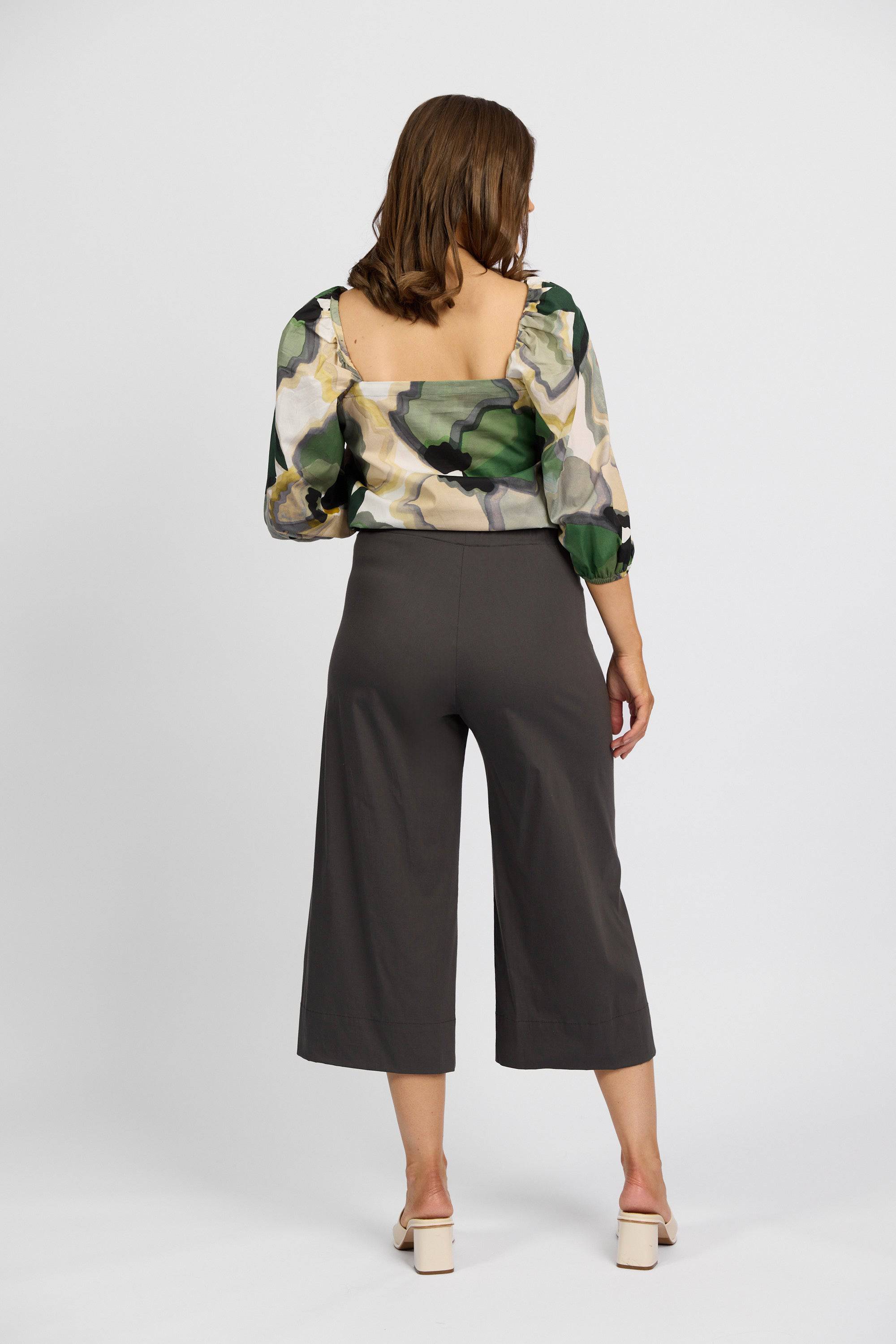 A woman faces away from the camera, wearing a sheer patterned blouse, Vassalli Paris Wide Leg Crop - Beluga pants in dark gray stretch fabric, and light heeled sandals against a plain background.
