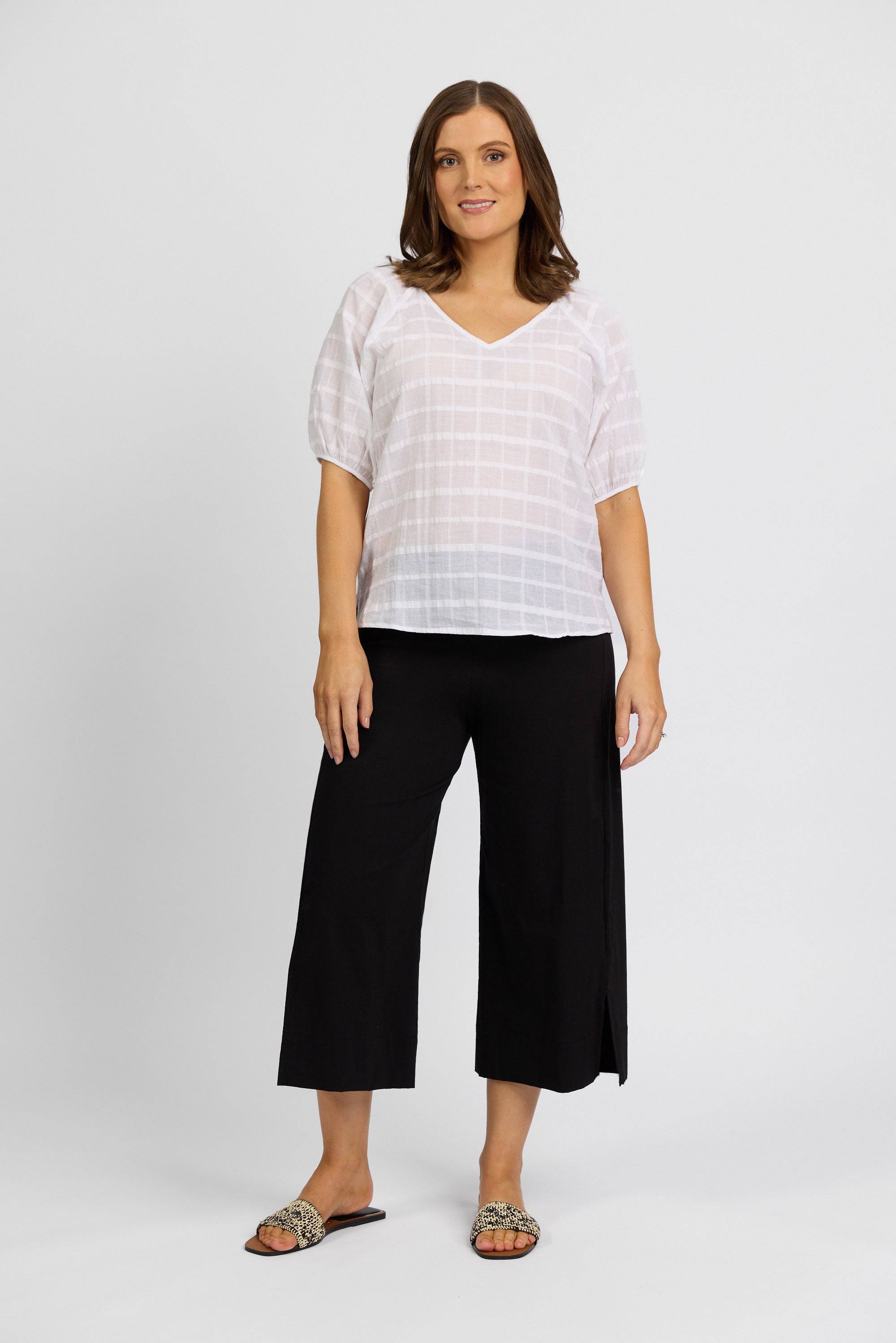 A woman with shoulder-length brown hair and a relaxed expression wears a loose white short-sleeve blouse, Vassalli Paris Wide Leg Crop - Black pants, and leopard-print slide sandals against a plain background.