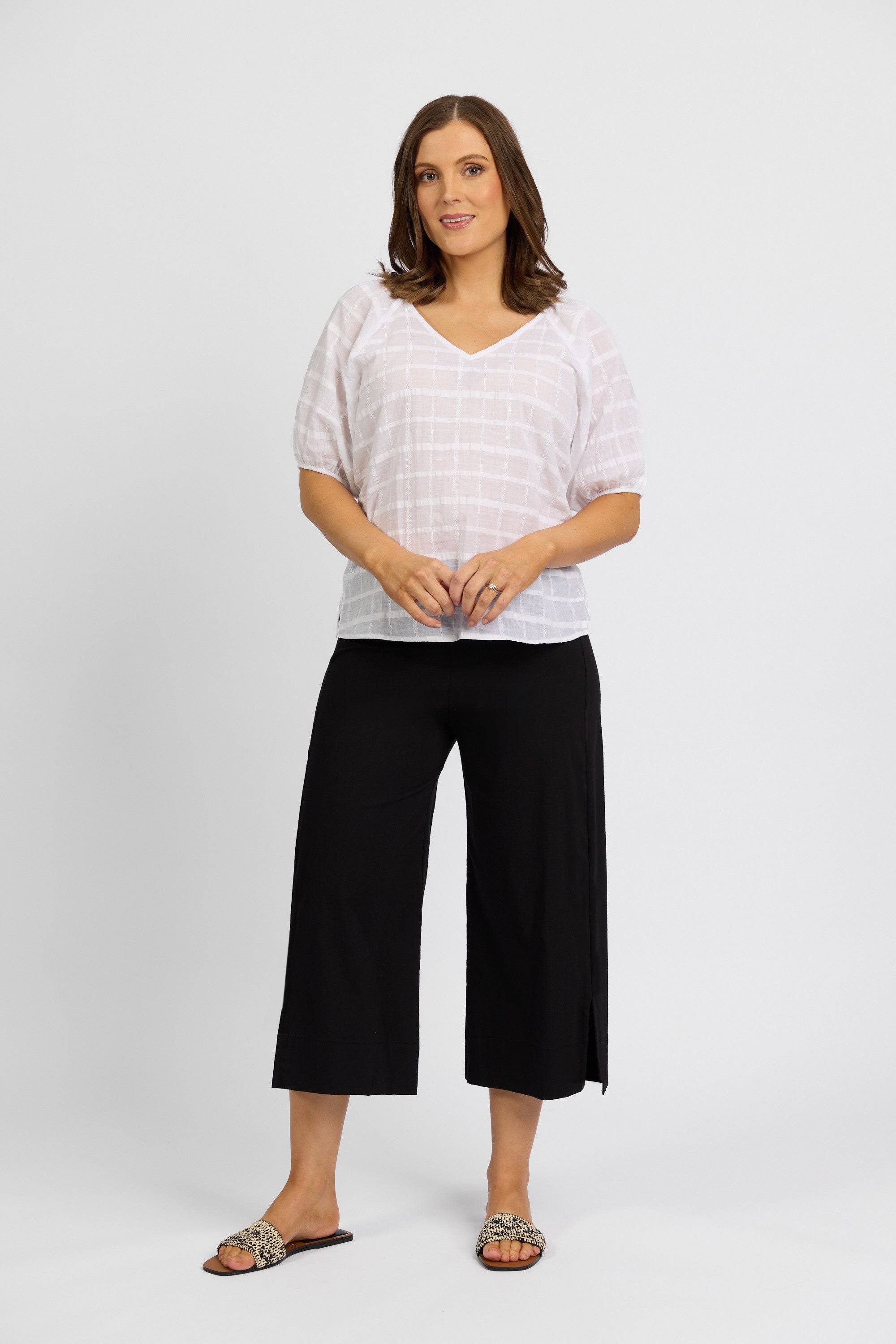 A woman stands against a white background, wearing a loose white short-sleeve blouse, Vassalli Paris Wide Leg Crop - Black pants in stretch fabric, and flat embellished strap slip-on sandals.