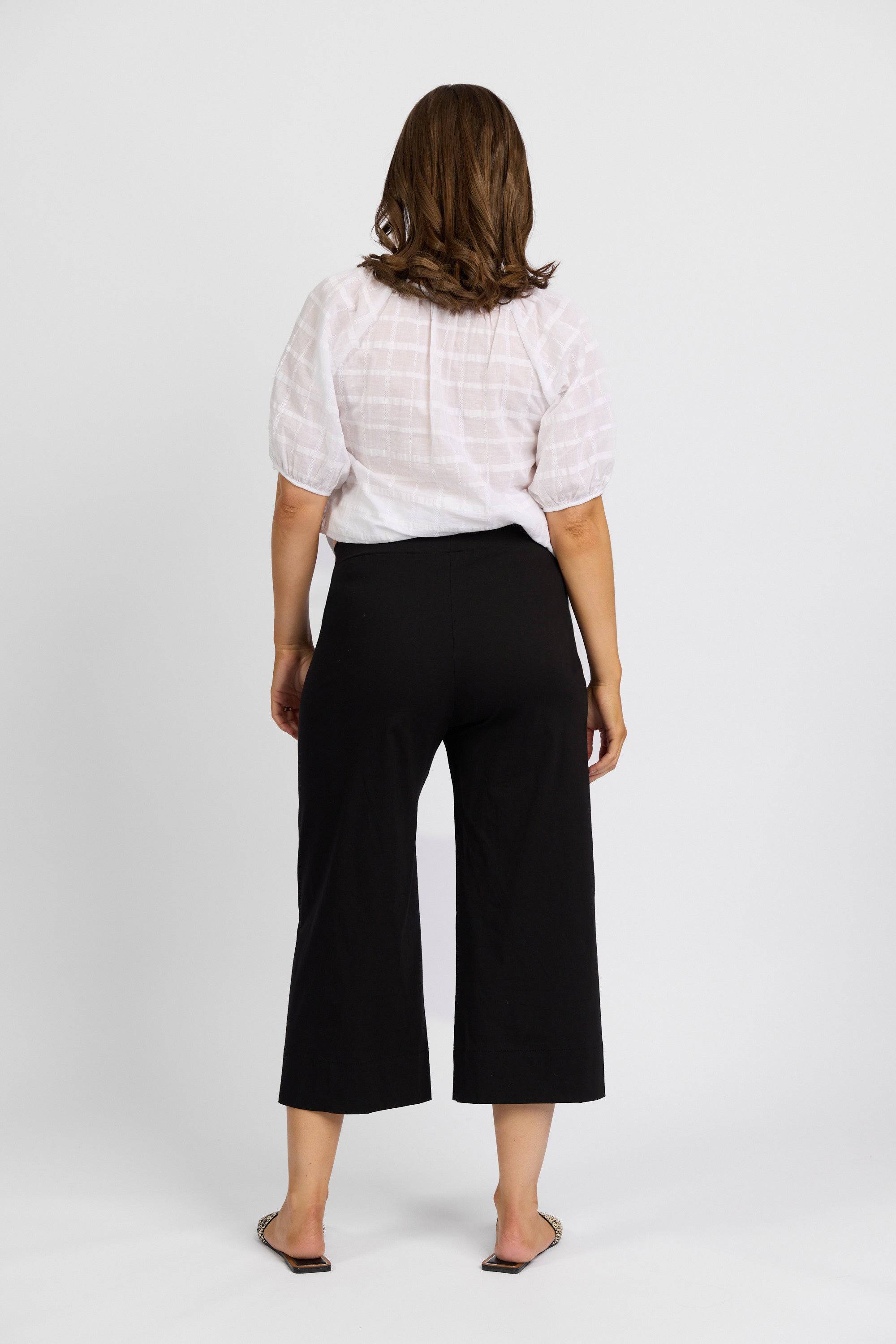 A woman with shoulder-length brown hair stands facing away, wearing a white short-sleeved blouse and Vassalli Paris Wide Leg Crop - Black pants, made from stretch fabric in New Zealand, with flat shoes against a plain white background.