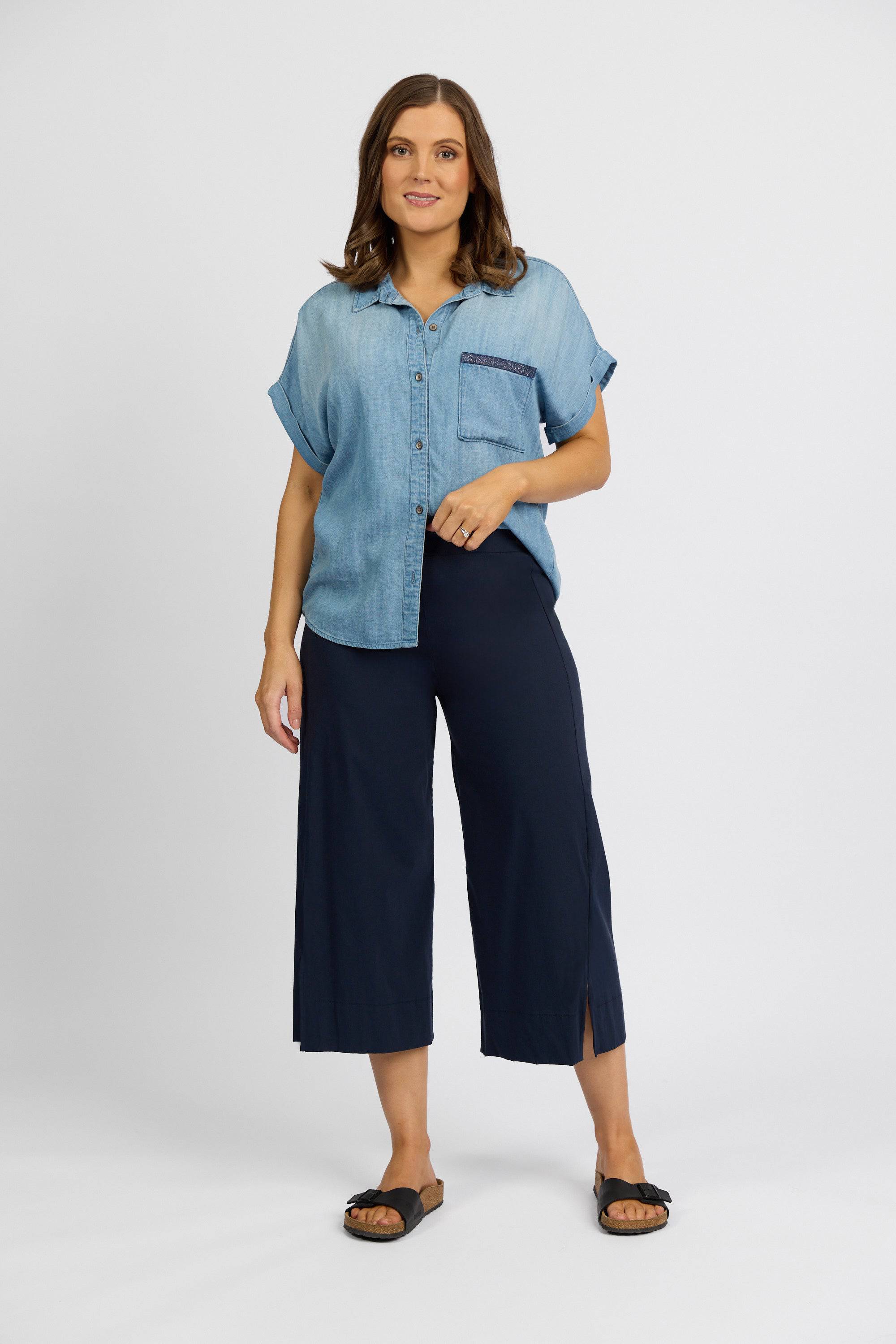A woman with shoulder-length brown hair stands against a white background, wearing a short-sleeve denim button-up, Vassalli's Paris Wide Leg Crop - Inka pants in stretch fabric, and black sandals. She smiles slightly.