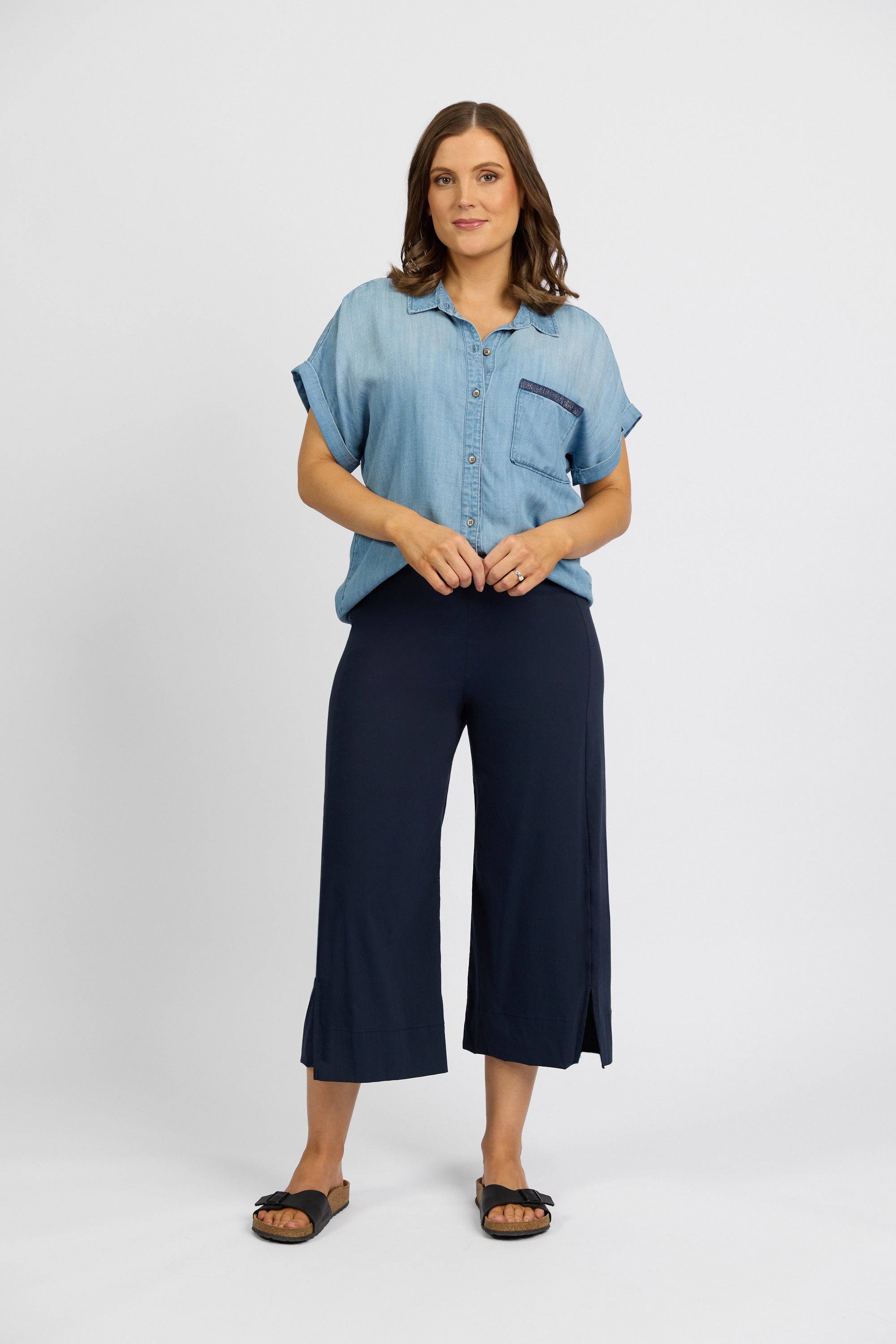 A woman stands against a white background wearing a short-sleeve light denim button-up shirt, black slide sandals, and Vassalli's Paris Wide Leg Crop - Inka pants. Made in New Zealand.