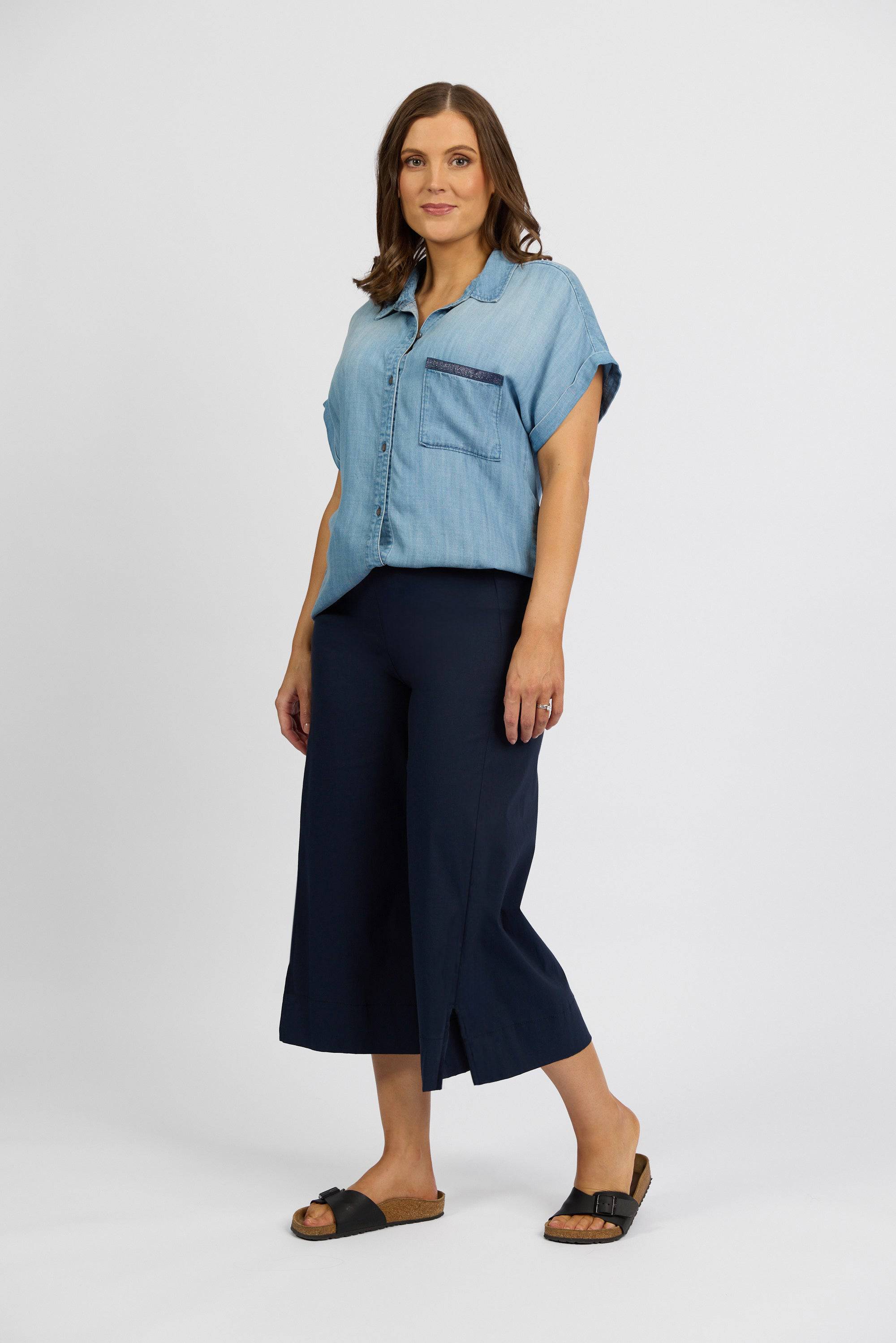 A woman with shoulder-length brown hair stands against a plain background, wearing a light blue short-sleeve button-up shirt and Vassalli Paris Wide Leg Crop - Inka pants in dark stretch fabric, paired with black sandals. Made in New Zealand.