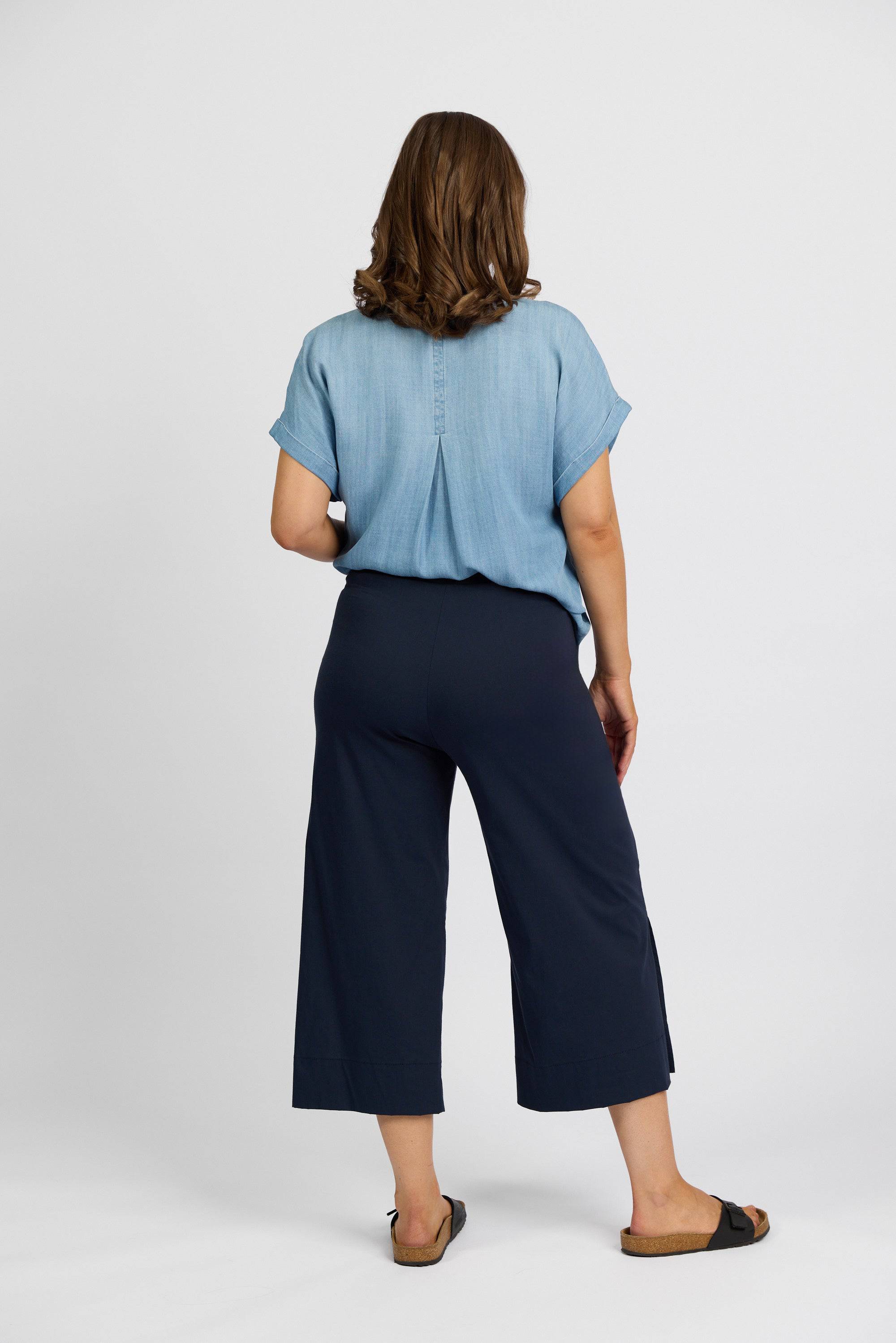 A woman with shoulder-length brown hair faces away, wearing a short-sleeve light denim shirt, Vassalli Paris Wide Leg Crop - Inka pants, and black sandals against a plain white background.