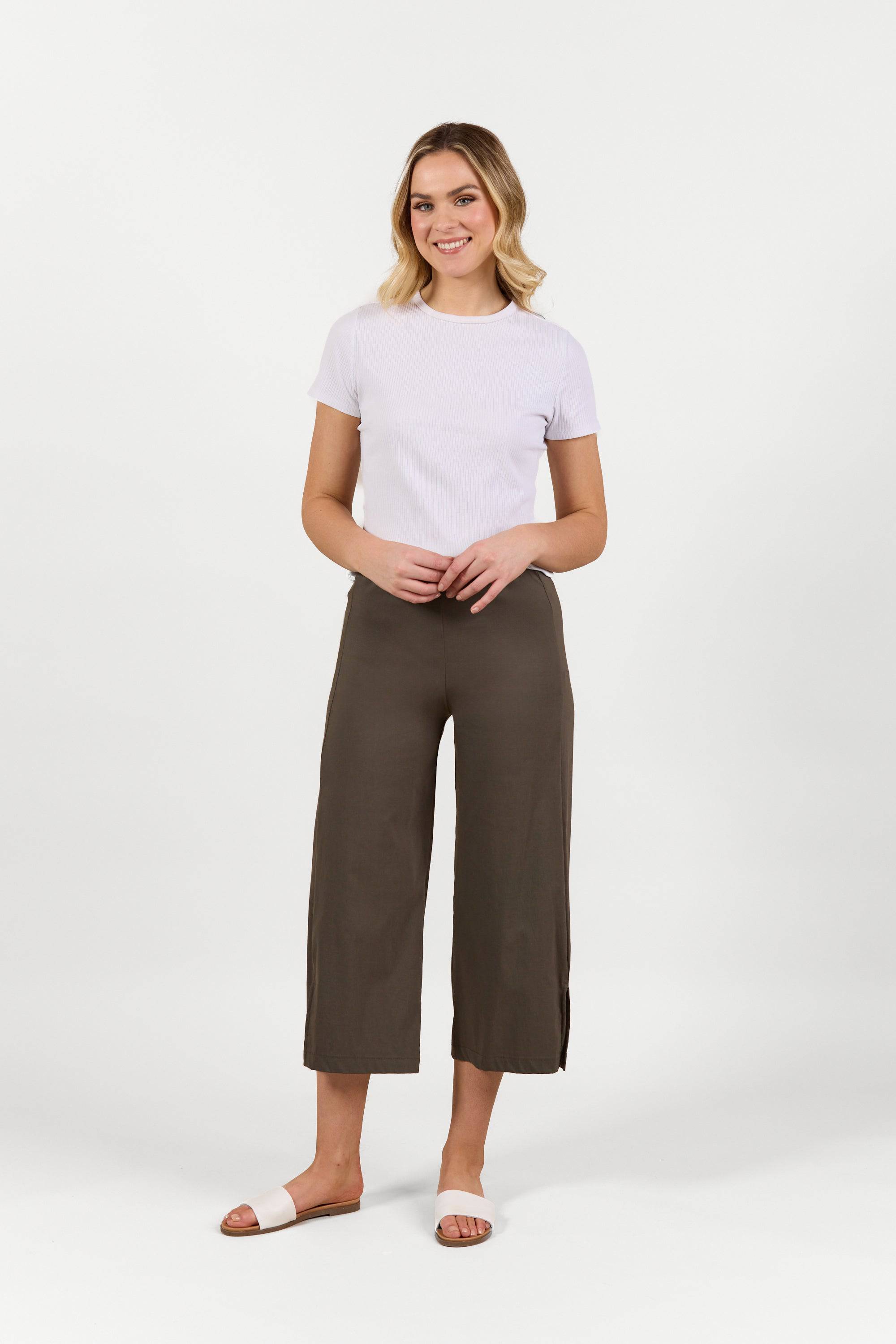 A woman with light hair smiles against a white background, wearing a short-sleeve white top and Vassalli Paris Wide Leg Crop pants in khaki stretch fabric, paired with white slide sandals. Her hands are loosely clasped in front of her.