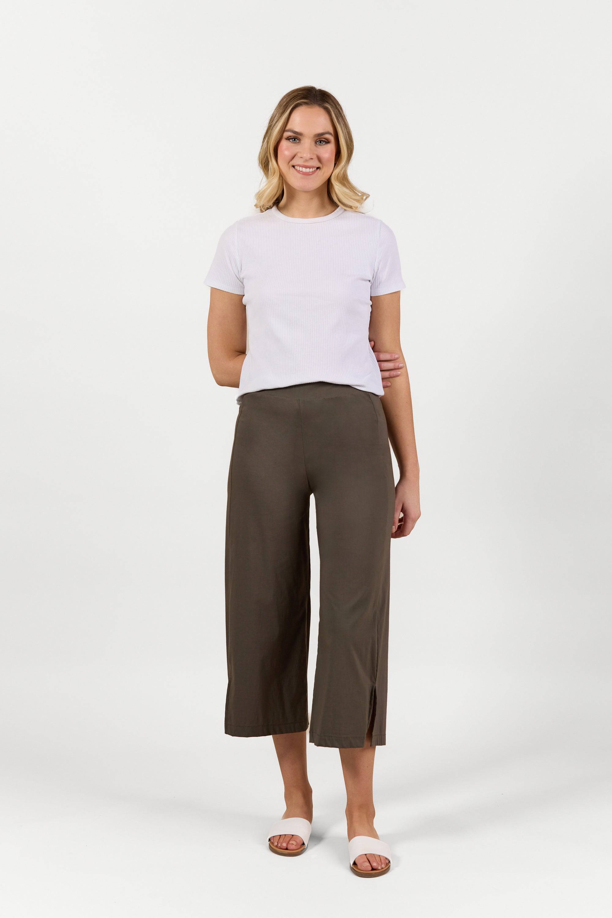 A woman stands facing forward, smiling, wearing a white short-sleeve t-shirt, Vassalli Paris Wide Leg Crop pants in khaki, and white sandals against a plain white background.