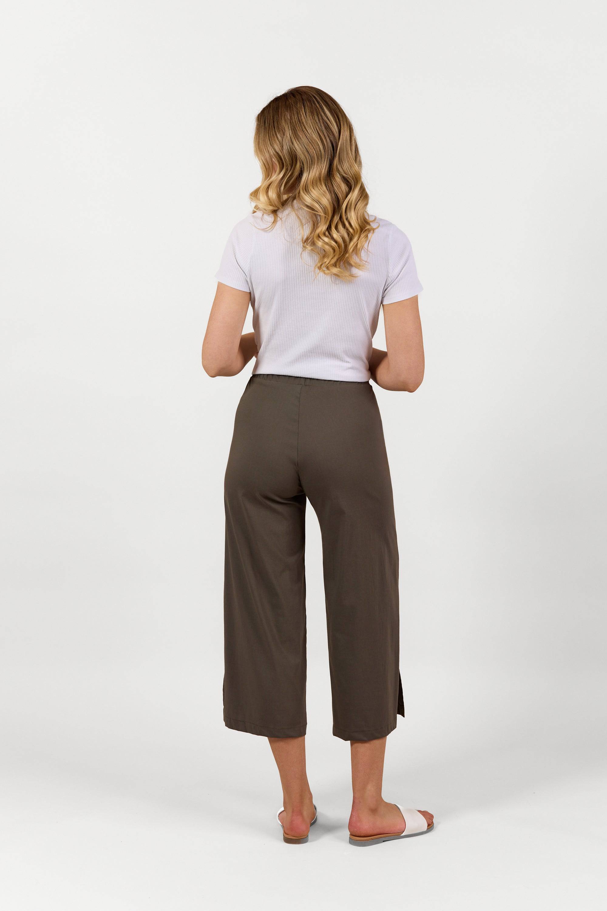 A woman with wavy blonde hair faces away, wearing a white short-sleeve shirt and Vassalli Paris Wide Leg Crop pants in Khaki, styled with sandals against a plain white background.