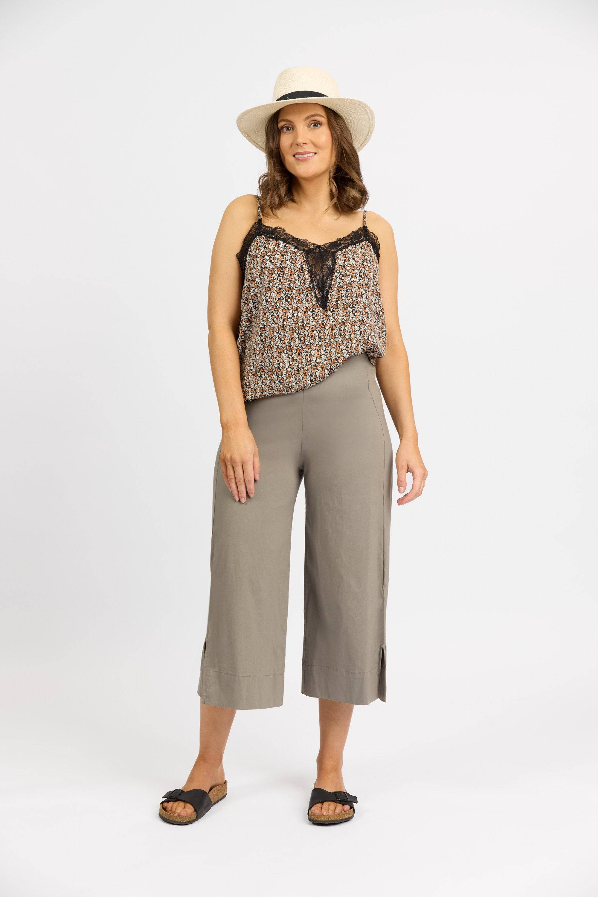A woman in a patterned sleeveless top, Vassalli Paris Wide Leg Crop - Latte pants, black sandals, and a white sunhat stands against a white background, looking forward and smiling slightly. Pants made in New Zealand.