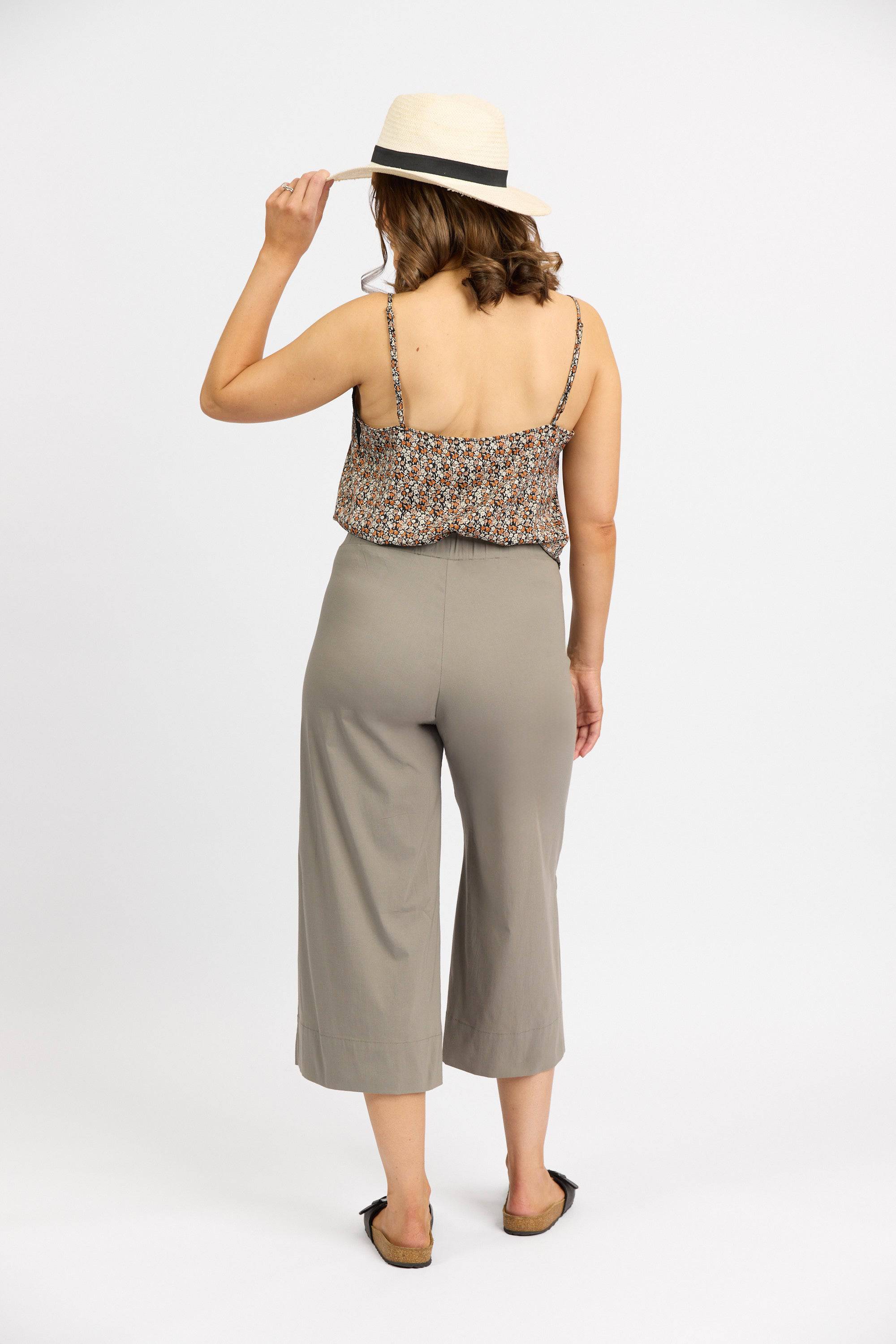 A woman stands facing away against a plain white background, wearing a straw hat, patterned sleeveless top, Vassalli Paris Wide Leg Crop – Latte pants in stretch fabric, and flat sandals, with her left hand touching her hat’s brim.