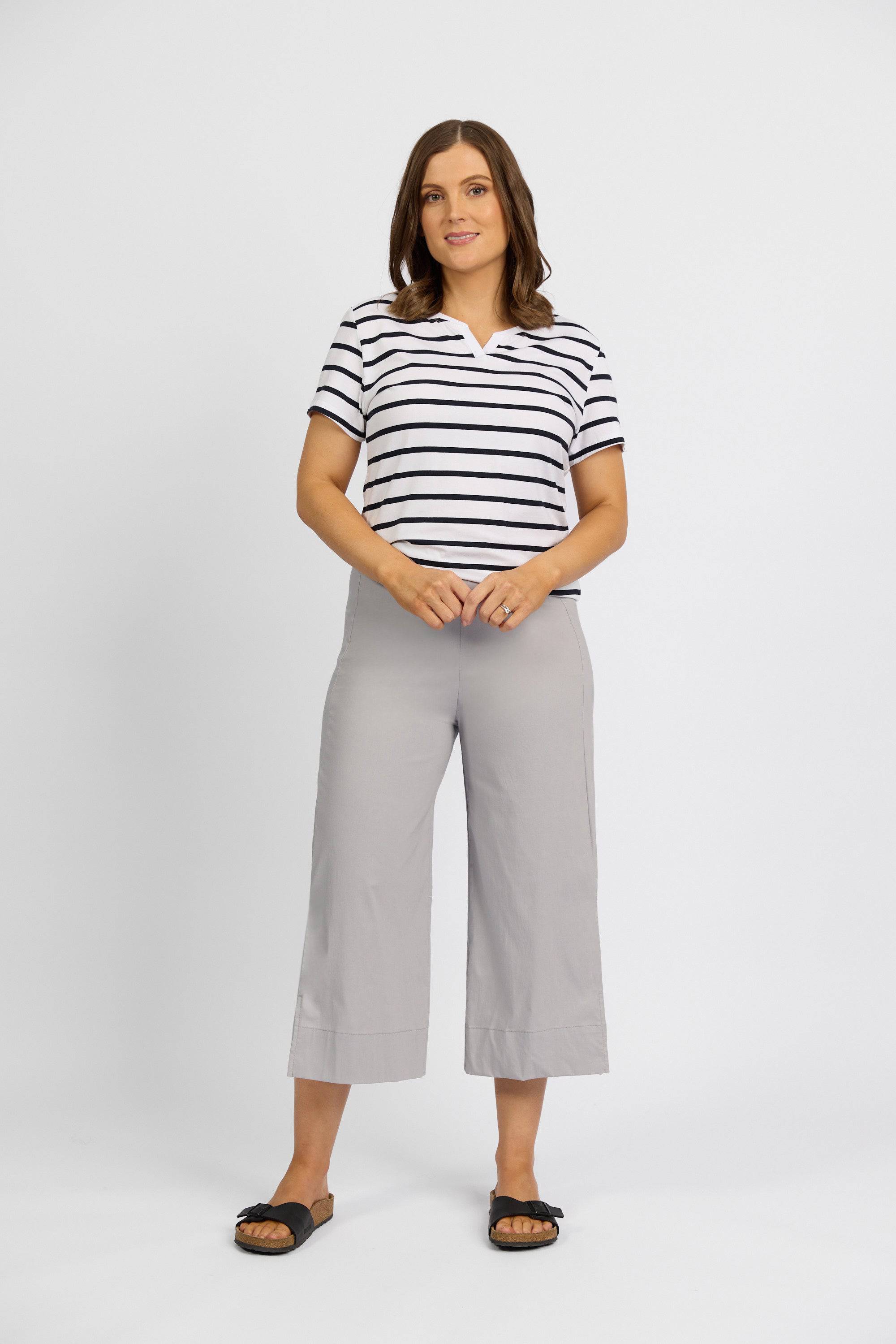 A woman with shoulder-length brown hair and a neutral expression stands against a white background, wearing a short-sleeve black-and-white striped top and Vassalli Paris Wide Leg Crop - Silver pants, paired with black slide sandals.