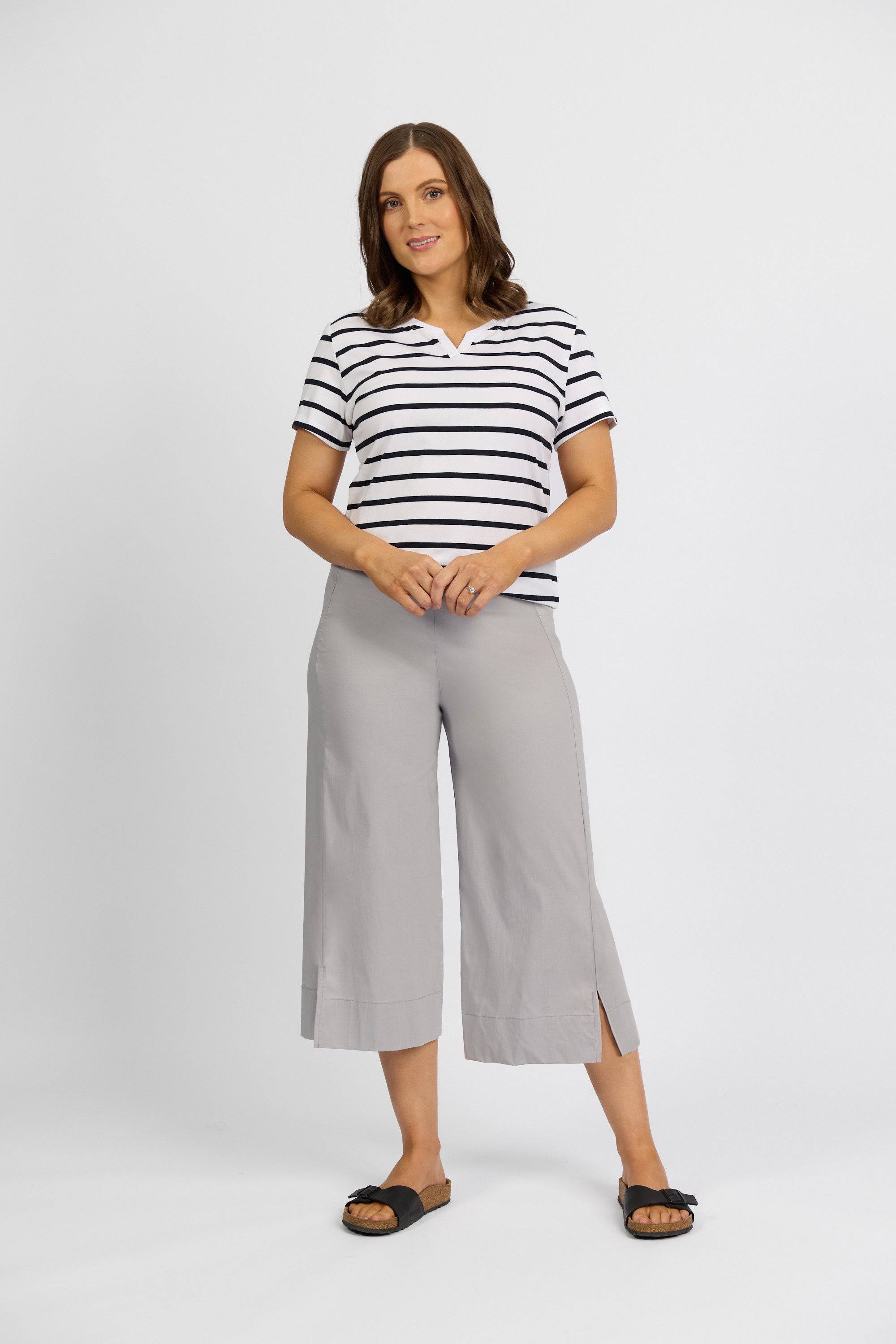 A woman stands against a white background, wearing a black and white striped short-sleeve top, Vassalli Paris Wide Leg Crop - Silver pants in stretch fabric with small side slits, and black slide sandals.