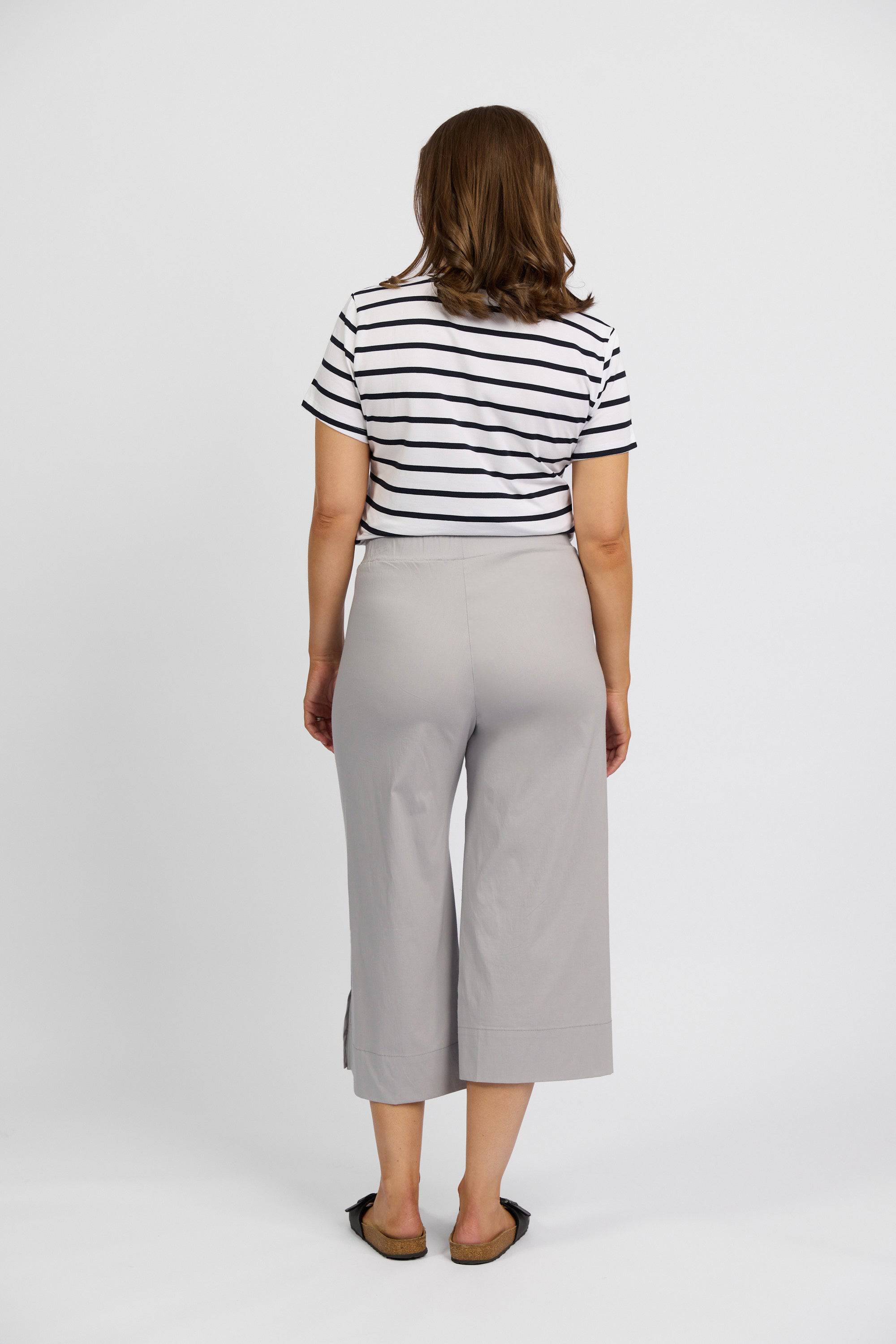 A woman faces away, wearing a short-sleeve black and white striped shirt, Vassalli Paris Wide Leg Crop - Silver pants, and black flats against a plain white background.