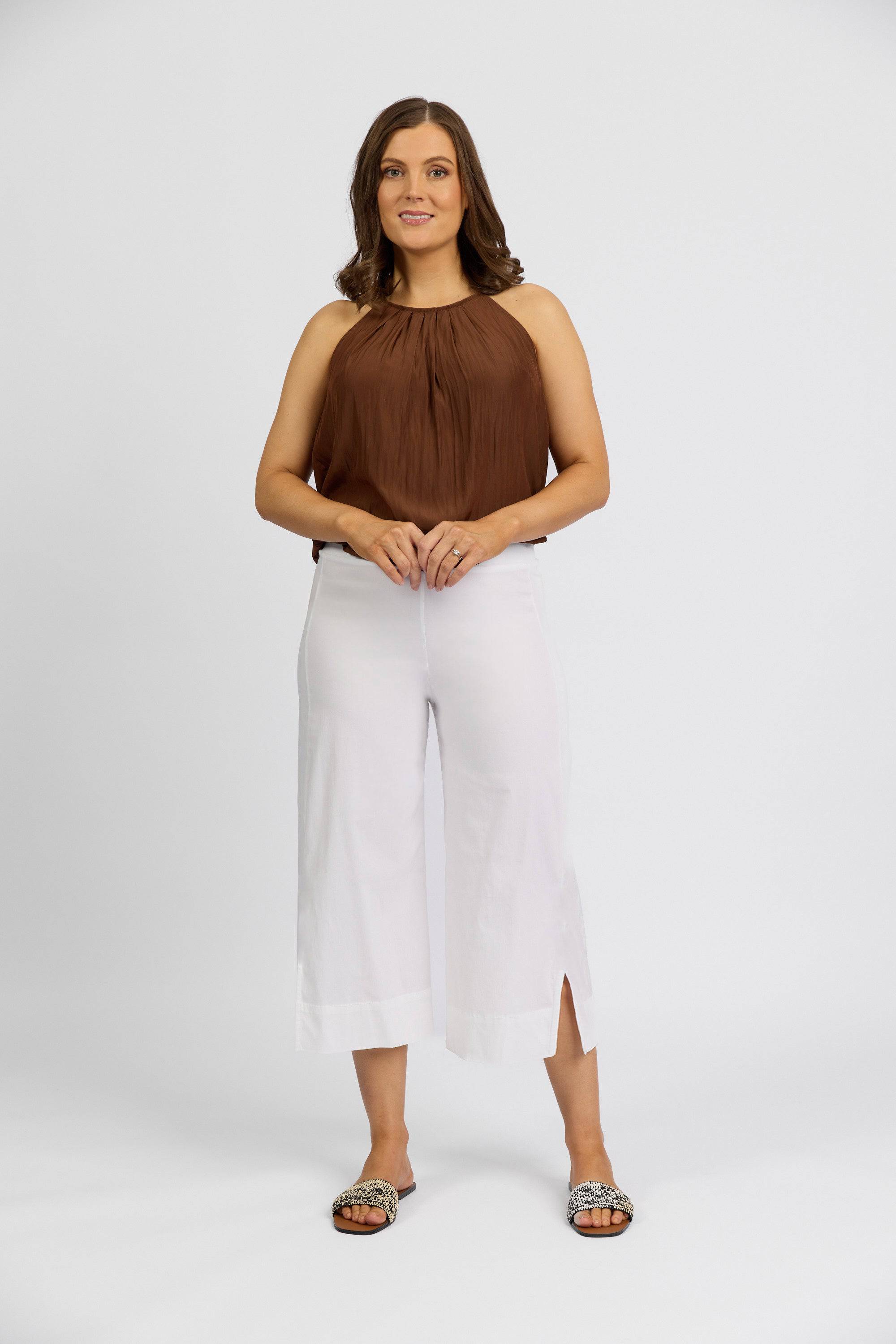 A woman stands against a white background, smiling with hands clasped, wearing a sleeveless brown halter top, Vassalli Paris Wide Leg Crop - White pants, and patterned flat sandals.