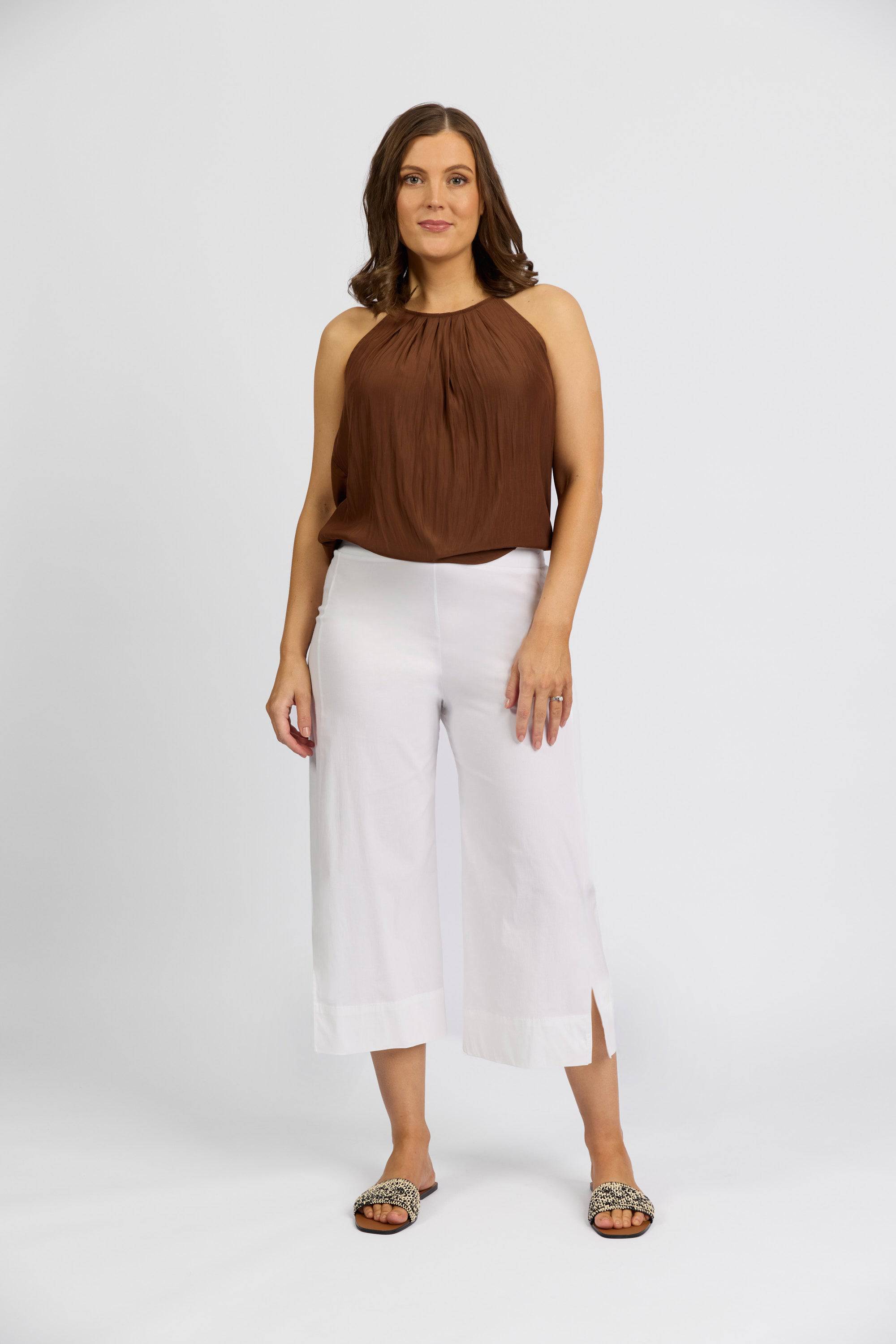 A woman stands against a plain background, wearing a sleeveless brown top with Vassalli Paris Wide Leg Crop - White pants made in New Zealand from stretch fabric, paired with open-toed sandals. Her shoulder-length brown hair frames her confident look.