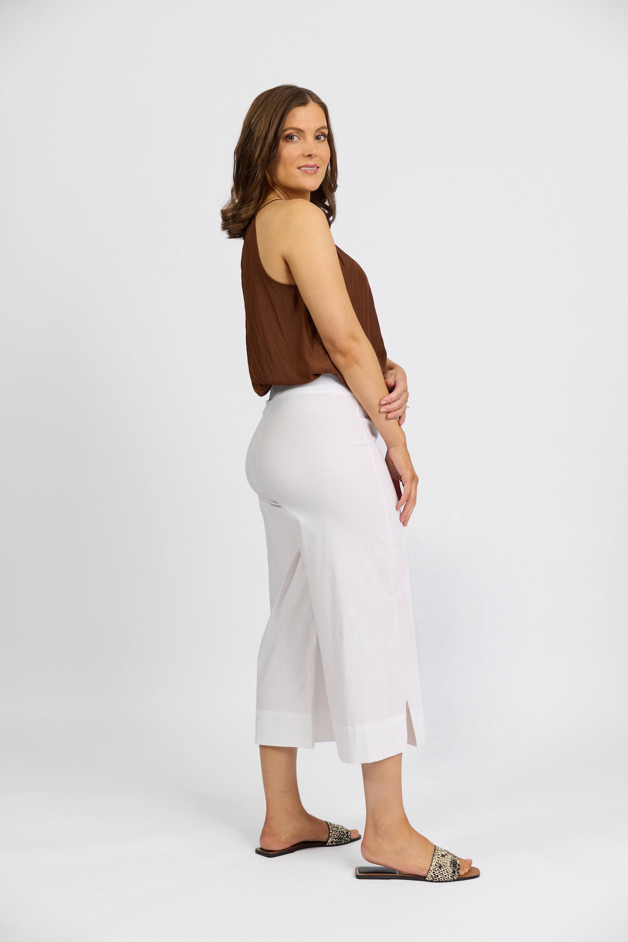 A woman stands against a white background, smiling with one hand on her arm. She wears a sleeveless brown top, patterned slide sandals, and the Vassalli Paris Wide Leg Crop - White: stretch fabric, wide-leg cropped pants.