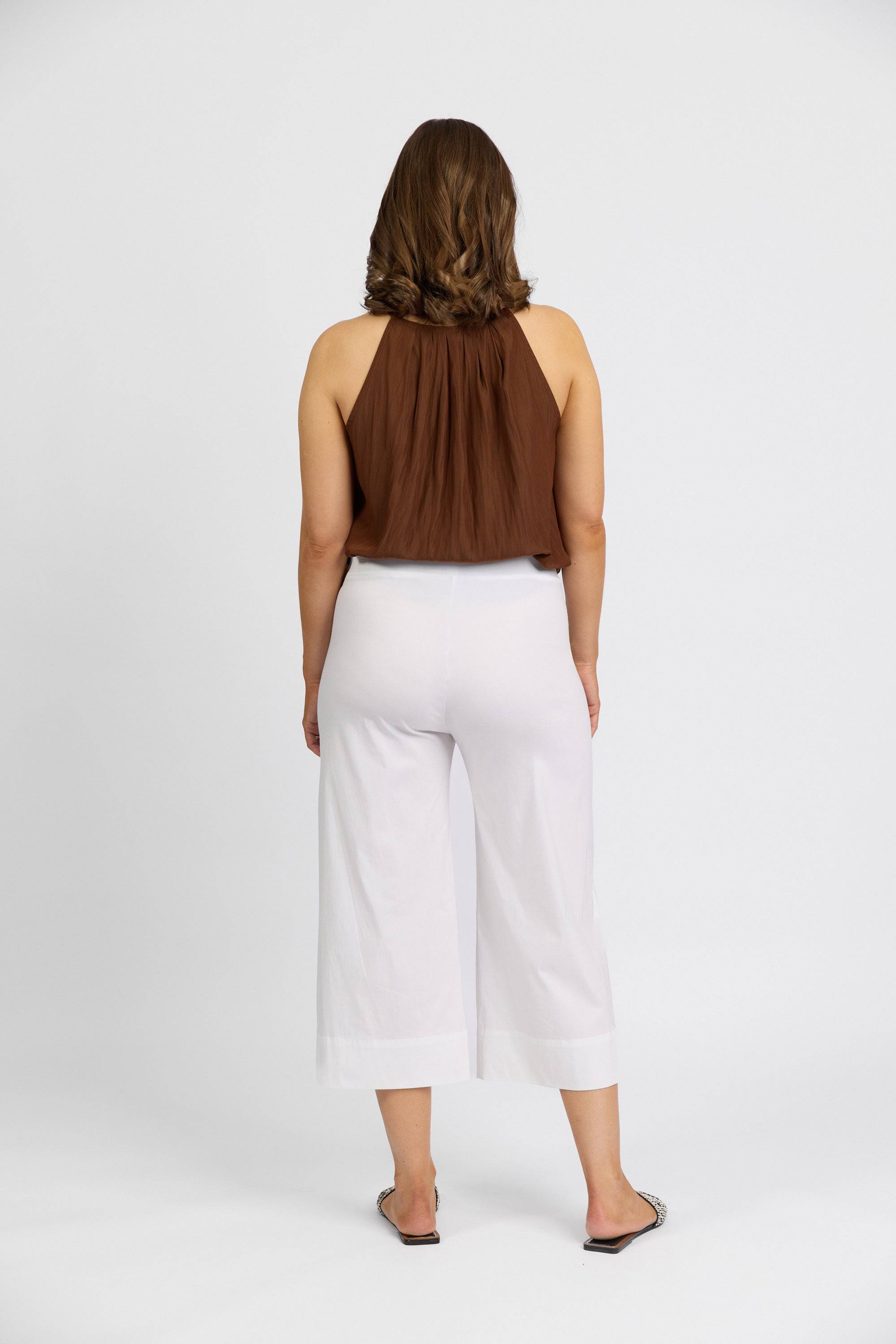 A woman, facing away from the camera, wears a sleeveless brown halter top, Vassalli Paris Wide Leg Crop - White pants, and flat shoes against a plain white background.