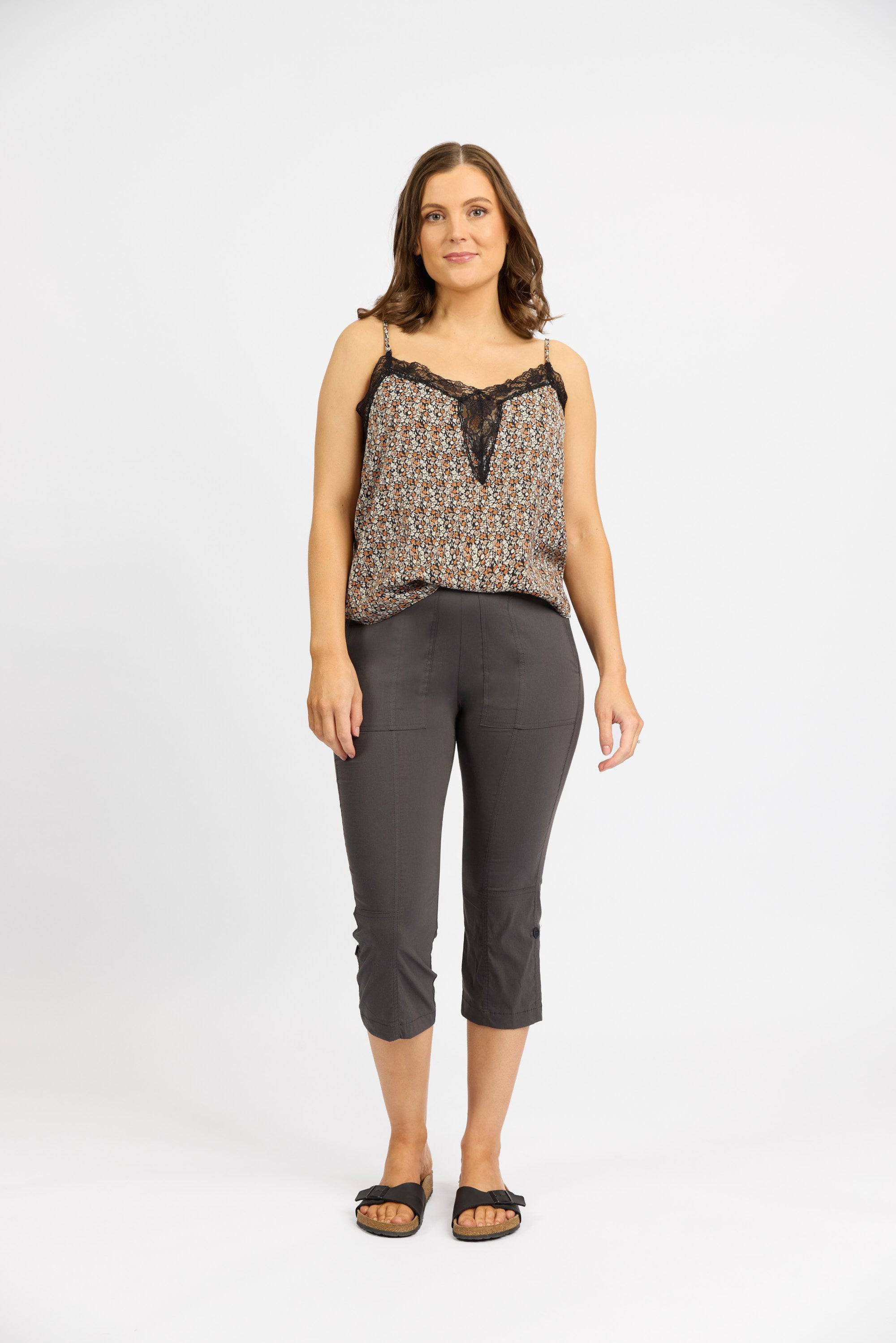 A woman with shoulder-length brown hair wears a patterned sleeveless top, Vassalli Paris 3/4 Cargo Short - Beluga made in New Zealand with stretch fabric, and black sandals, standing against a plain white background with a neutral expression.