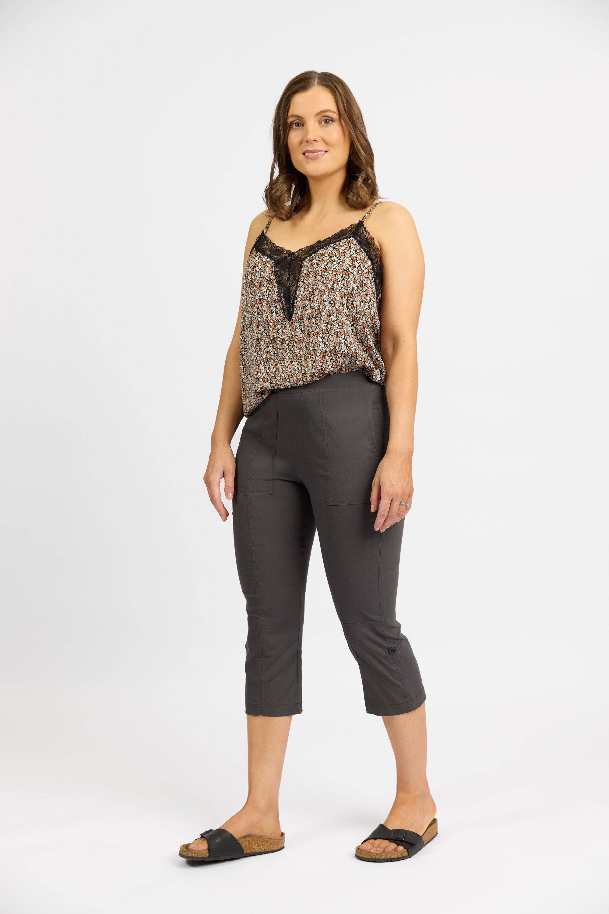 A woman with shoulder-length brown hair wears a sleeveless patterned top, Vassalli Paris 3/4 Cargo Short in Beluga, and black sandals, standing against a white background with a slight smile.