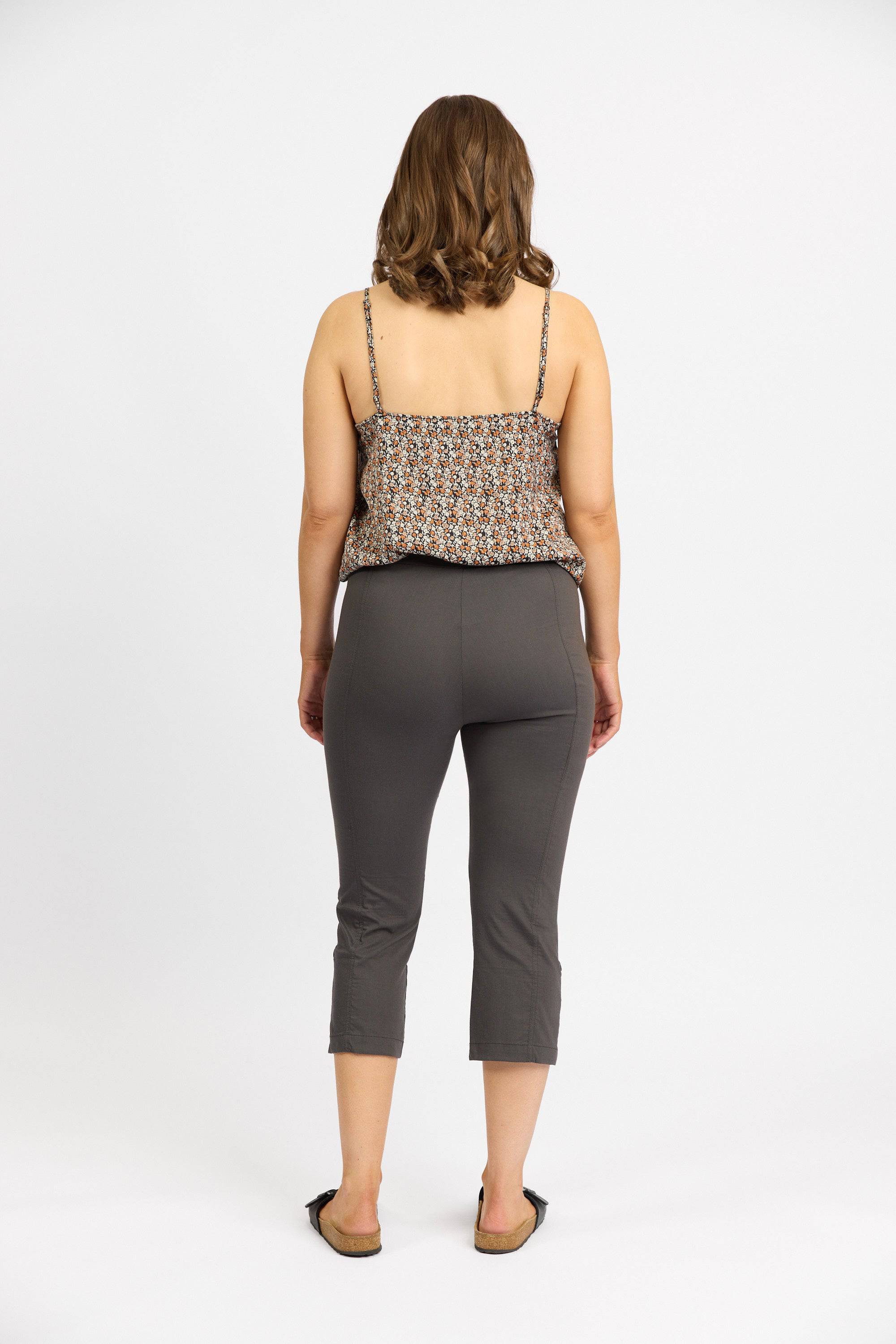 A woman with shoulder-length brown hair, facing away, wears a patterned sleeveless top, Vassalli Paris 3/4 Cargo Short - Beluga made in New Zealand from stretch fabric, and black flats against a plain white background.