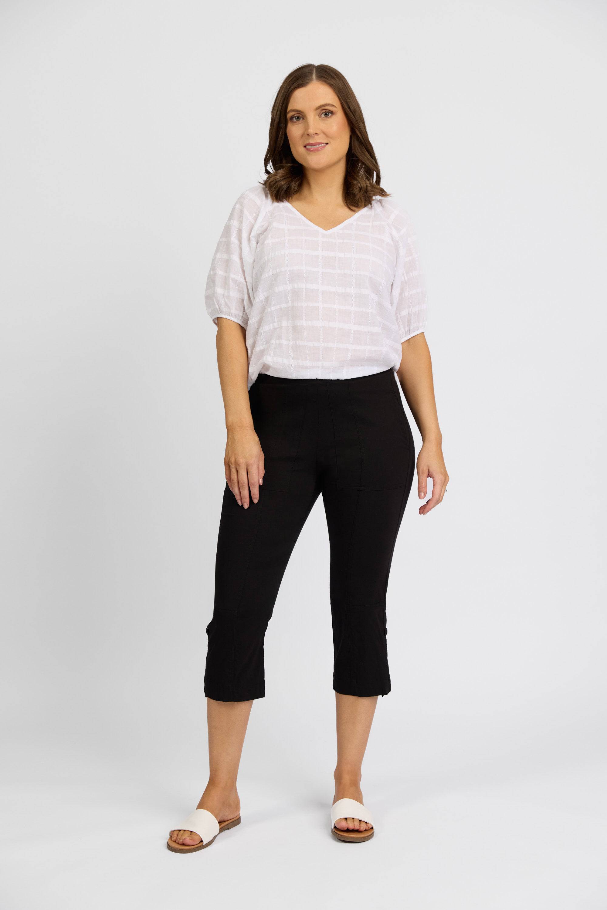 A woman with medium-length brown hair stands against a white background, wearing a white textured blouse and Vassalli Paris 3/4 Cargo Shorts in black, paired with white slide sandals. She looks at the camera with a neutral expression.