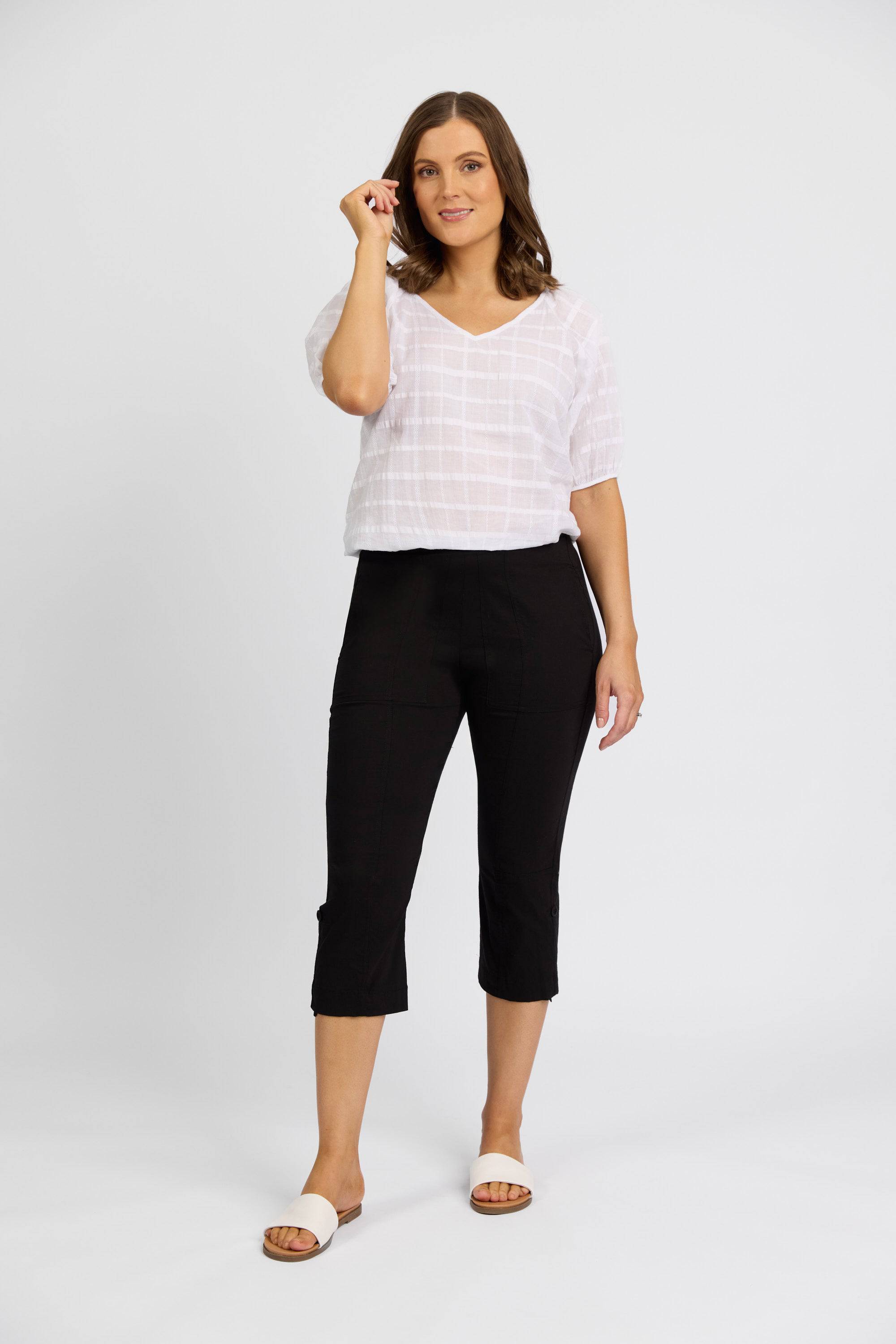 A woman with medium-length brown hair, smiling slightly, stands against a white background wearing a textured white short-sleeve blouse, beige slide sandals, and Vassalli Paris 3/4 Cargo Short in black.