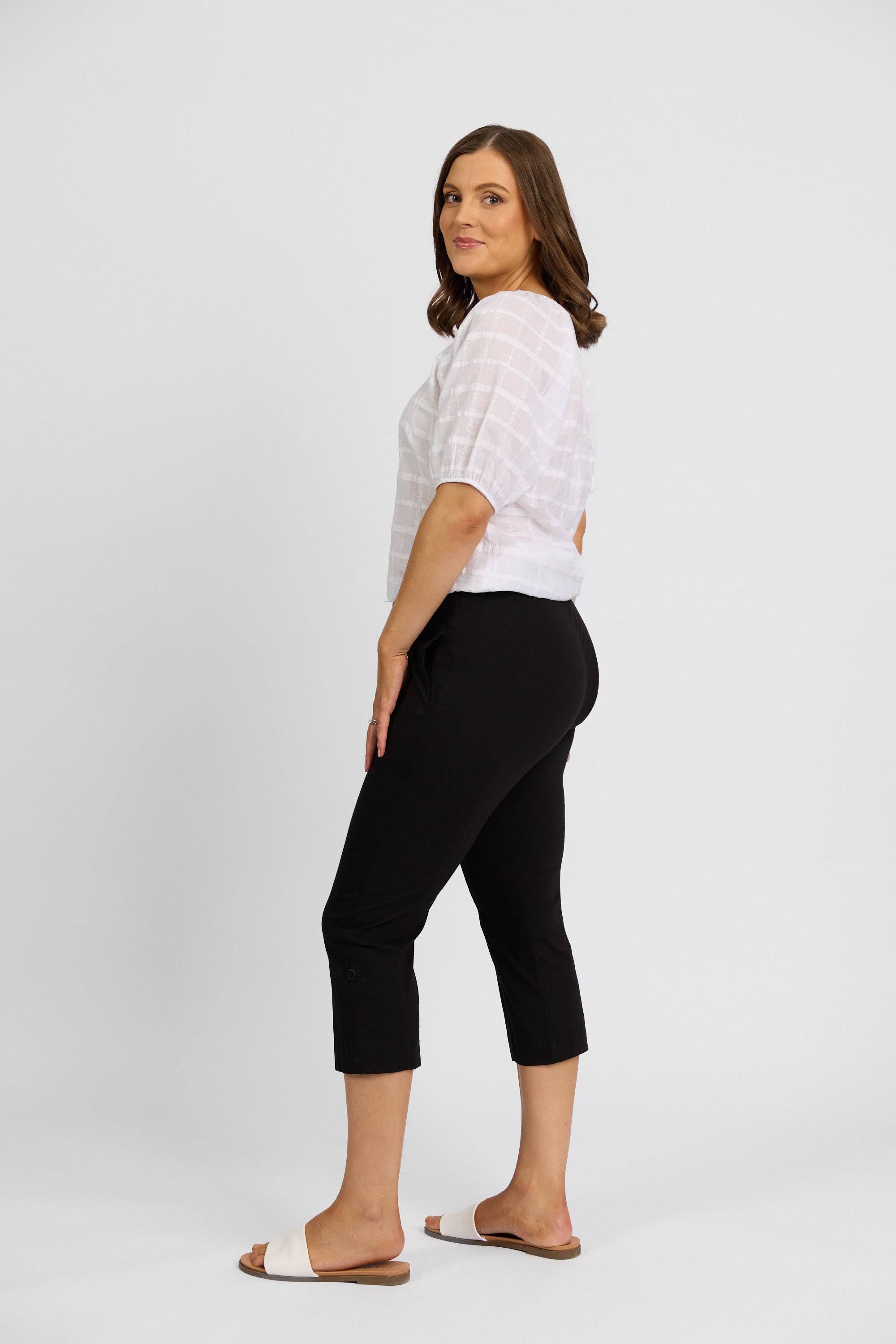 A woman with shoulder-length brown hair stands sideways, looking at the camera. She wears a white short-sleeve blouse, Vassalli Paris 3/4 Cargo Short in black, and white slide sandals against a plain white background.