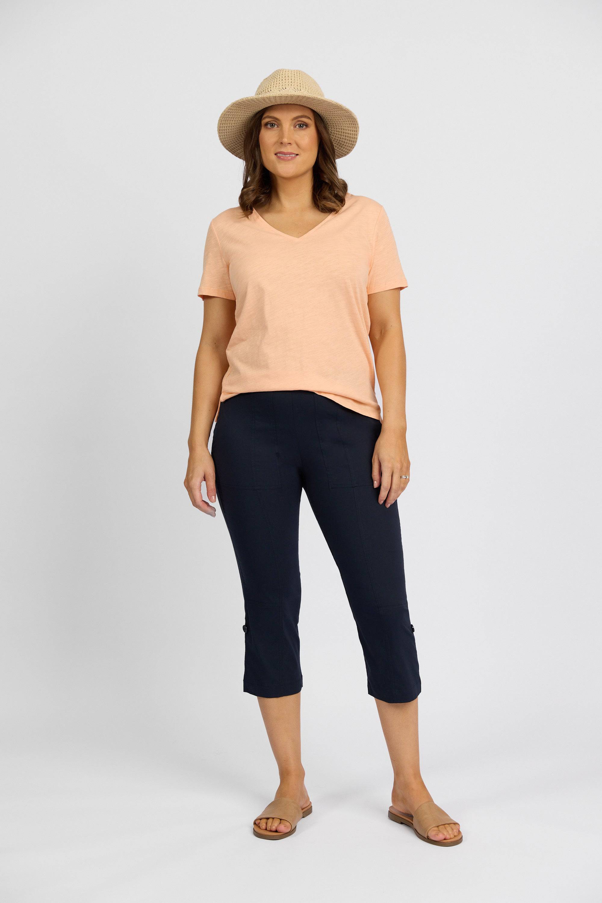A woman stands against a plain backdrop wearing a straw hat, peach V-neck tee, Vassalli Paris 3/4 Cargo Short - Inka in stretch fabric, and tan sandals. She faces forward with a relaxed expression and casual posture.