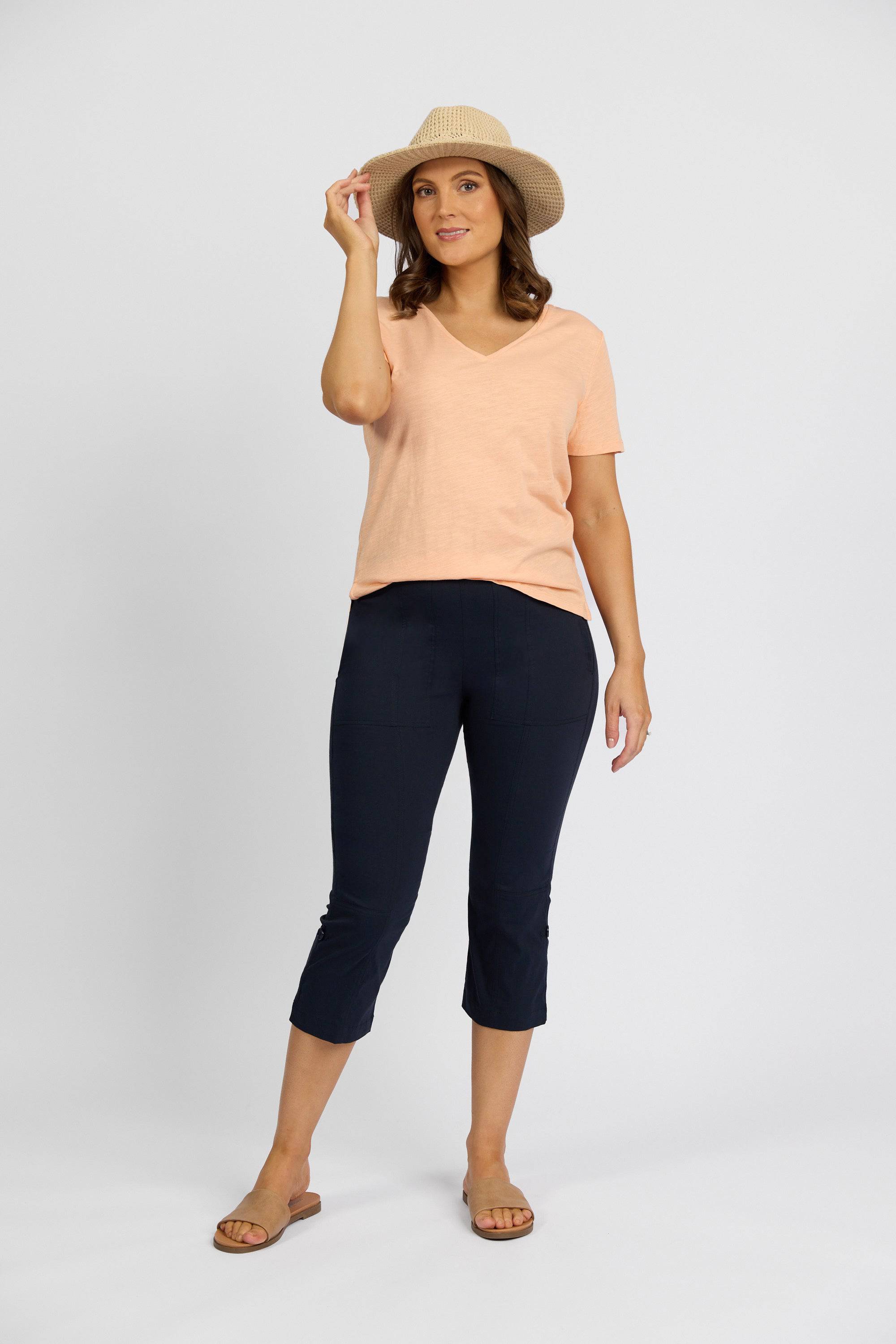 A woman smiles against a plain background, wearing a peach stretch V-neck tee, tan sandals, a beige sun hat, and Vassalli Paris 3/4 Cargo Shorts in Inka, holding her hat’s brim with one hand.