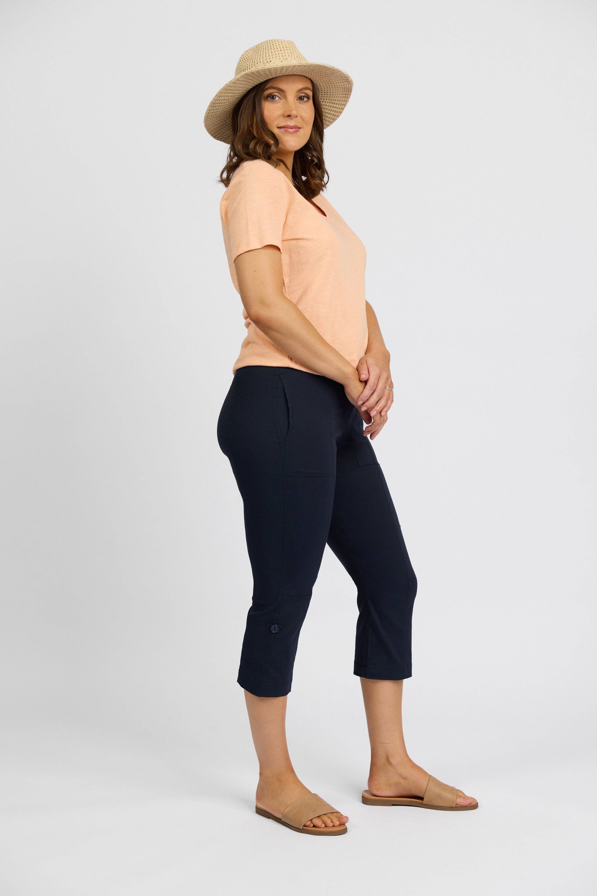A woman stands sideways in a relaxed pose, wearing a straw hat, peach short-sleeve shirt, Vassalli Paris 3/4 Cargo Short - Inka, and tan sandals against a plain white background.