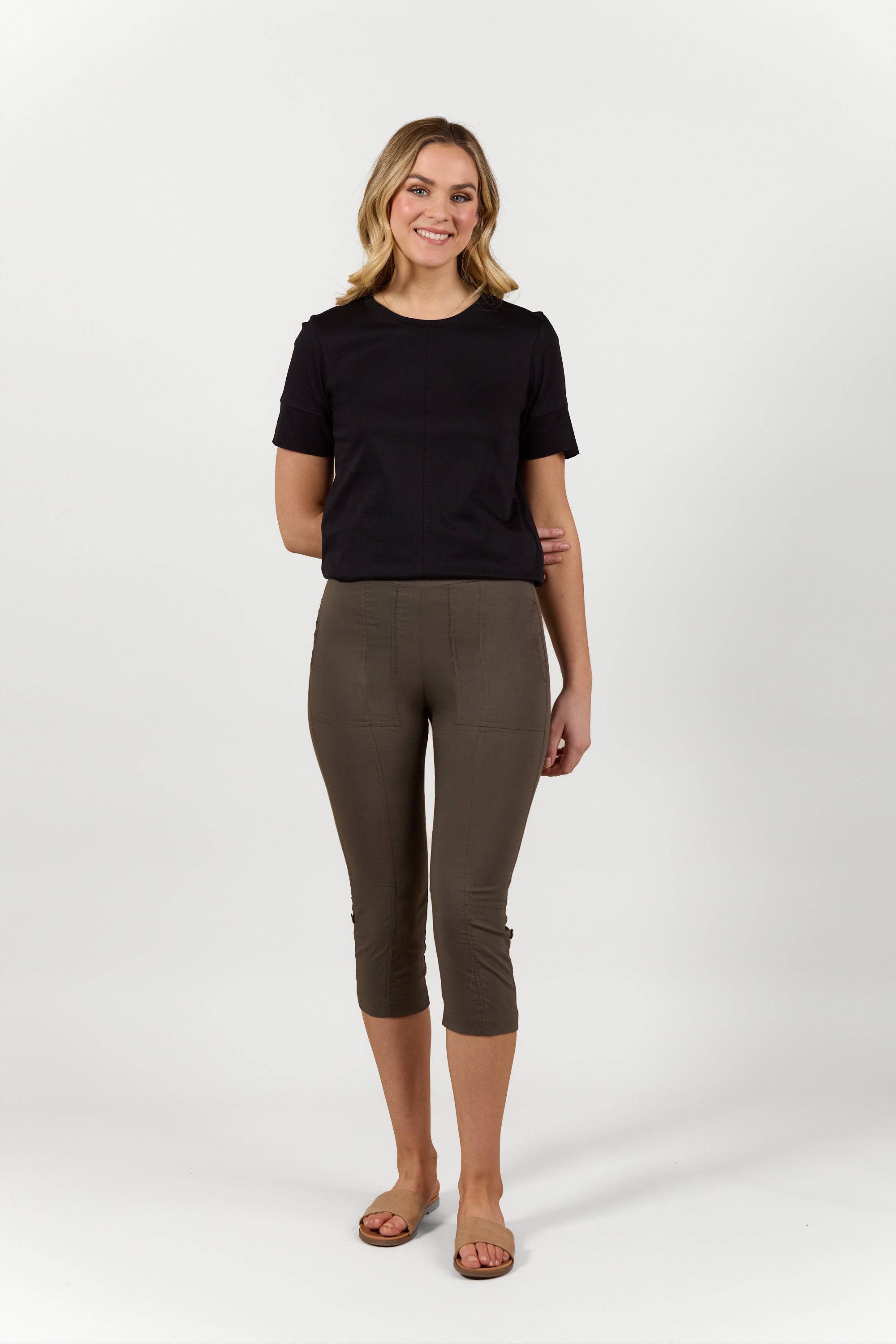 A woman with shoulder-length blonde hair stands smiling in a studio, wearing a black top, beige sandals, and Vassalli Paris 3/4 Cargo Short - Khaki made in New Zealand from stretch fabric. The background is plain white.