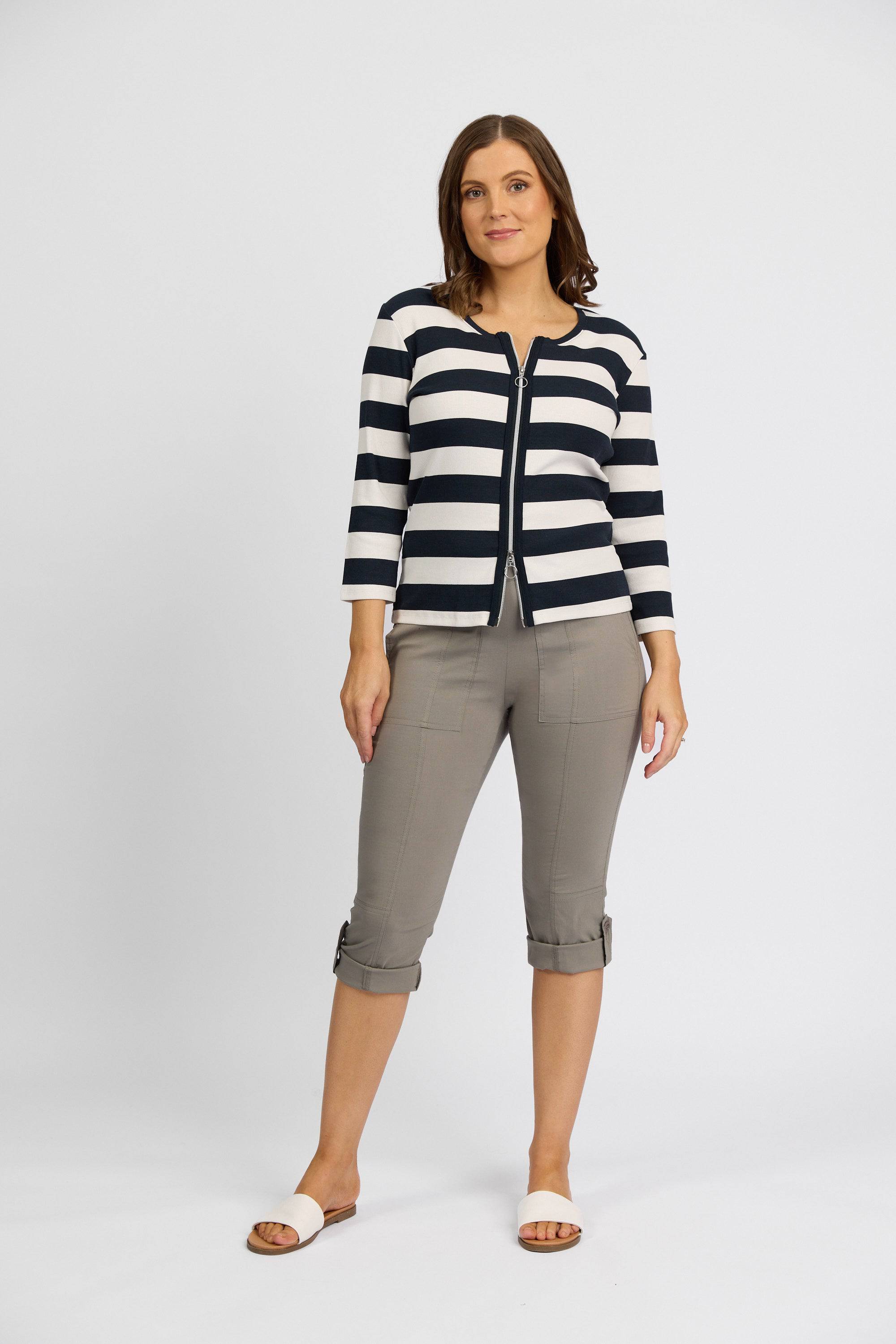 A woman with shoulder-length brown hair and a neutral expression stands against a white background, wearing a black and white striped cardigan, Vassalli Paris 3/4 Cargo Short in Latte, and white slide sandals.