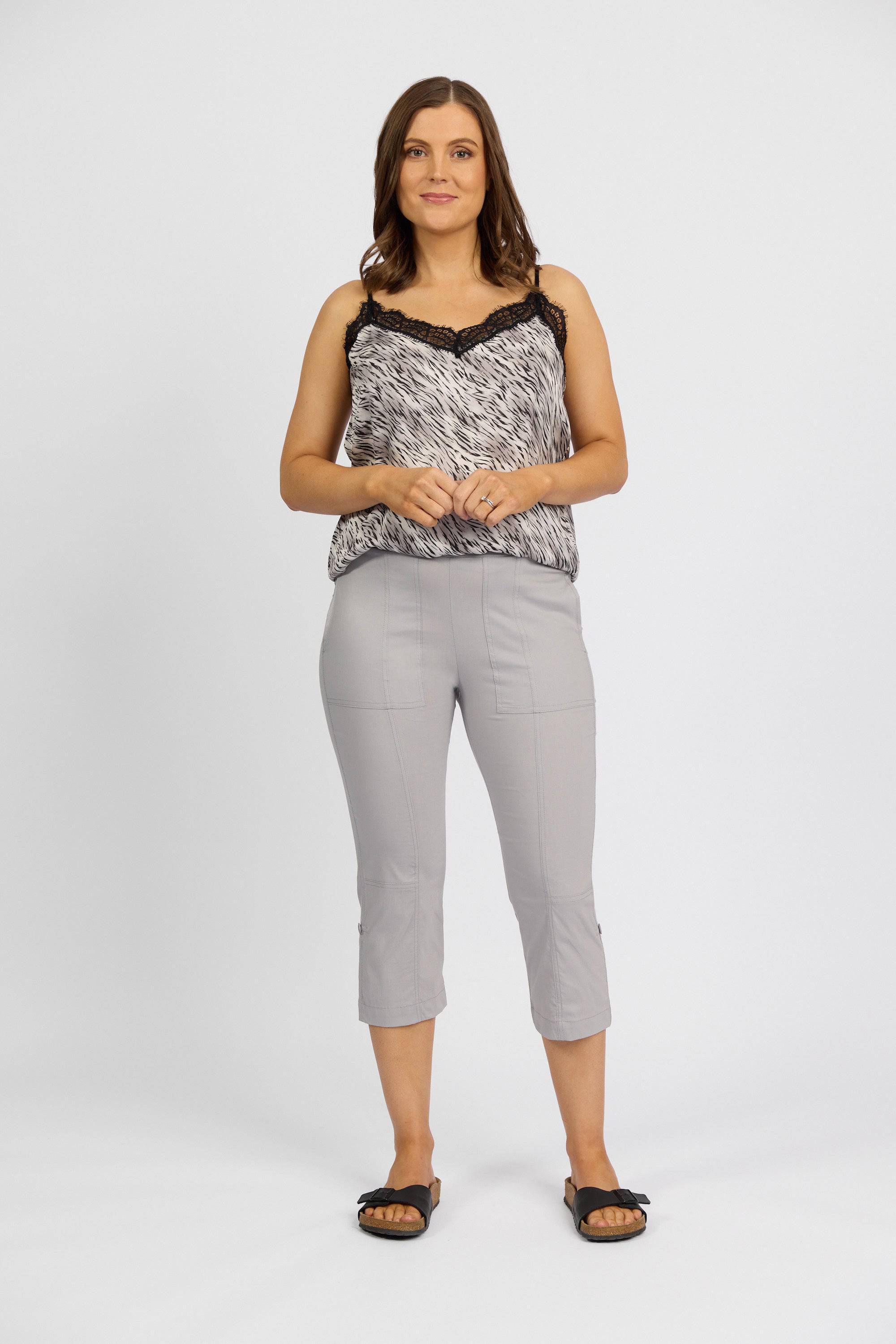 A woman stands against a white background, wearing a black-and-white patterned sleeveless top with lace trim and Vassalli Paris 3/4 Cargo Shorts in Silver. She smiles warmly, hands clasped, finishing the look with black slide sandals.