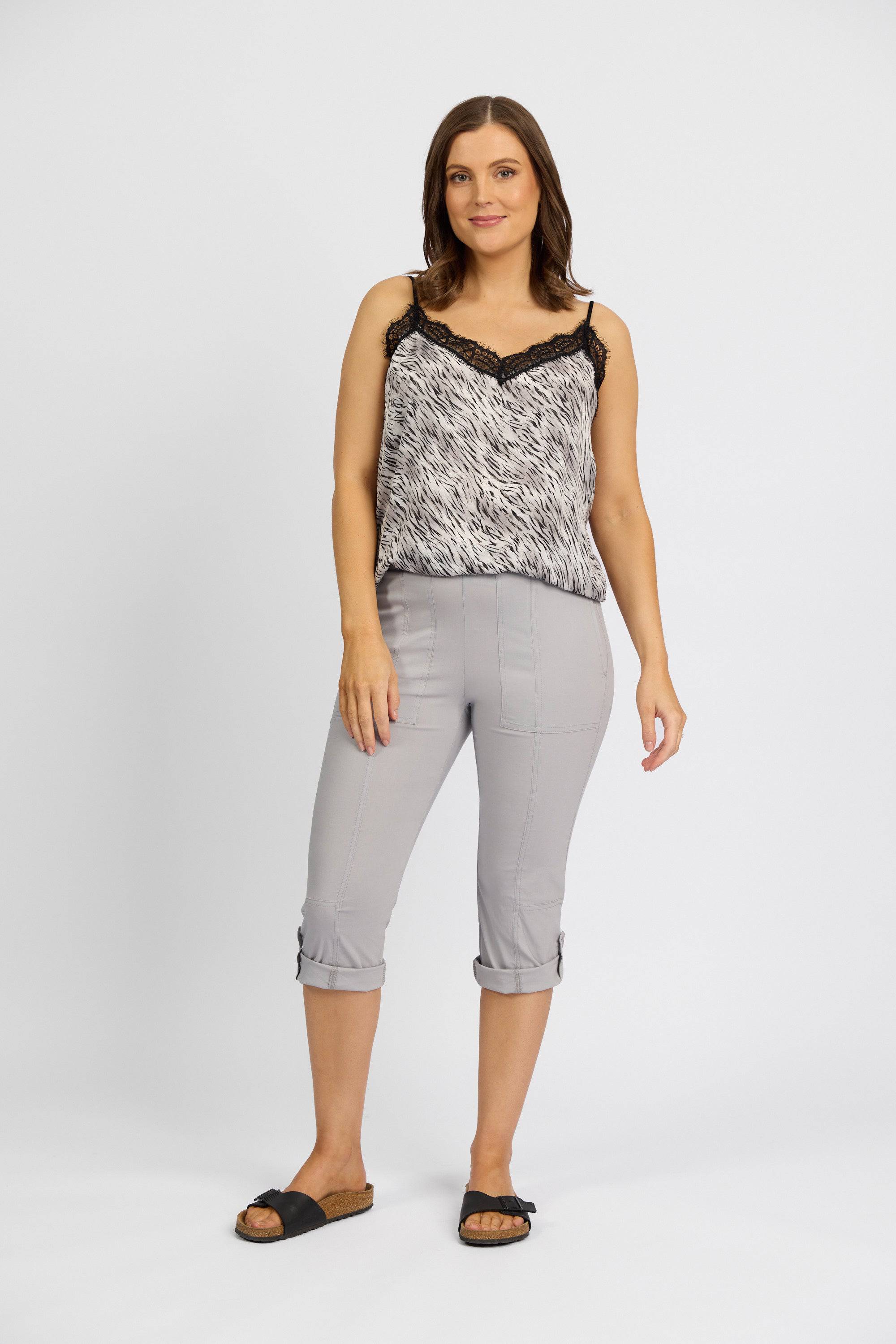 A woman smiles against a plain background, wearing a black and white patterned sleeveless top with black lace trim, Vassalli Paris 3/4 Cargo Short in Silver, and black slide sandals. One hand is relaxed by her side.