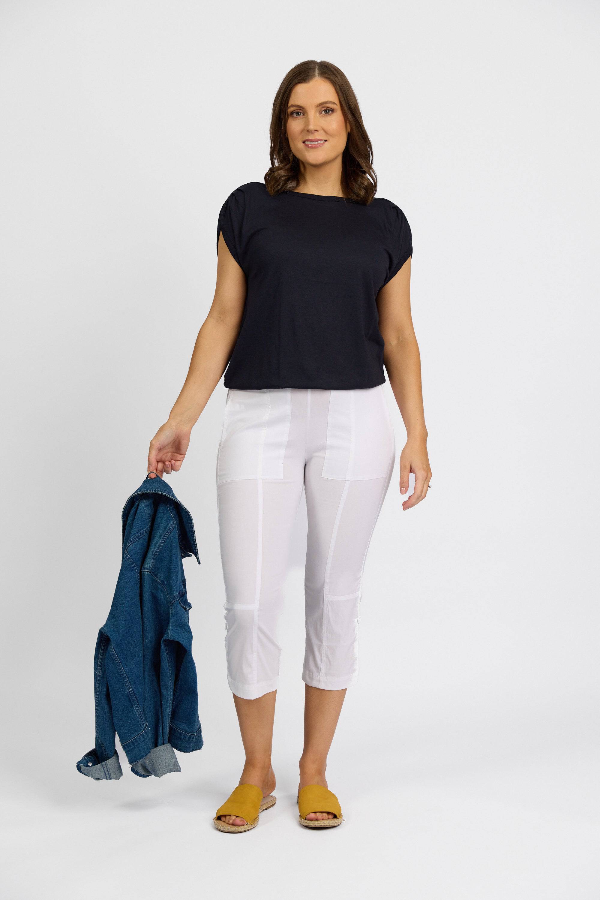 A woman stands against a white background wearing a black short-sleeve top, Vassalli Paris 3/4 Cargo Short in white, and yellow sandals. She holds a blue denim jacket in her left hand and looks relaxed and pleasant.