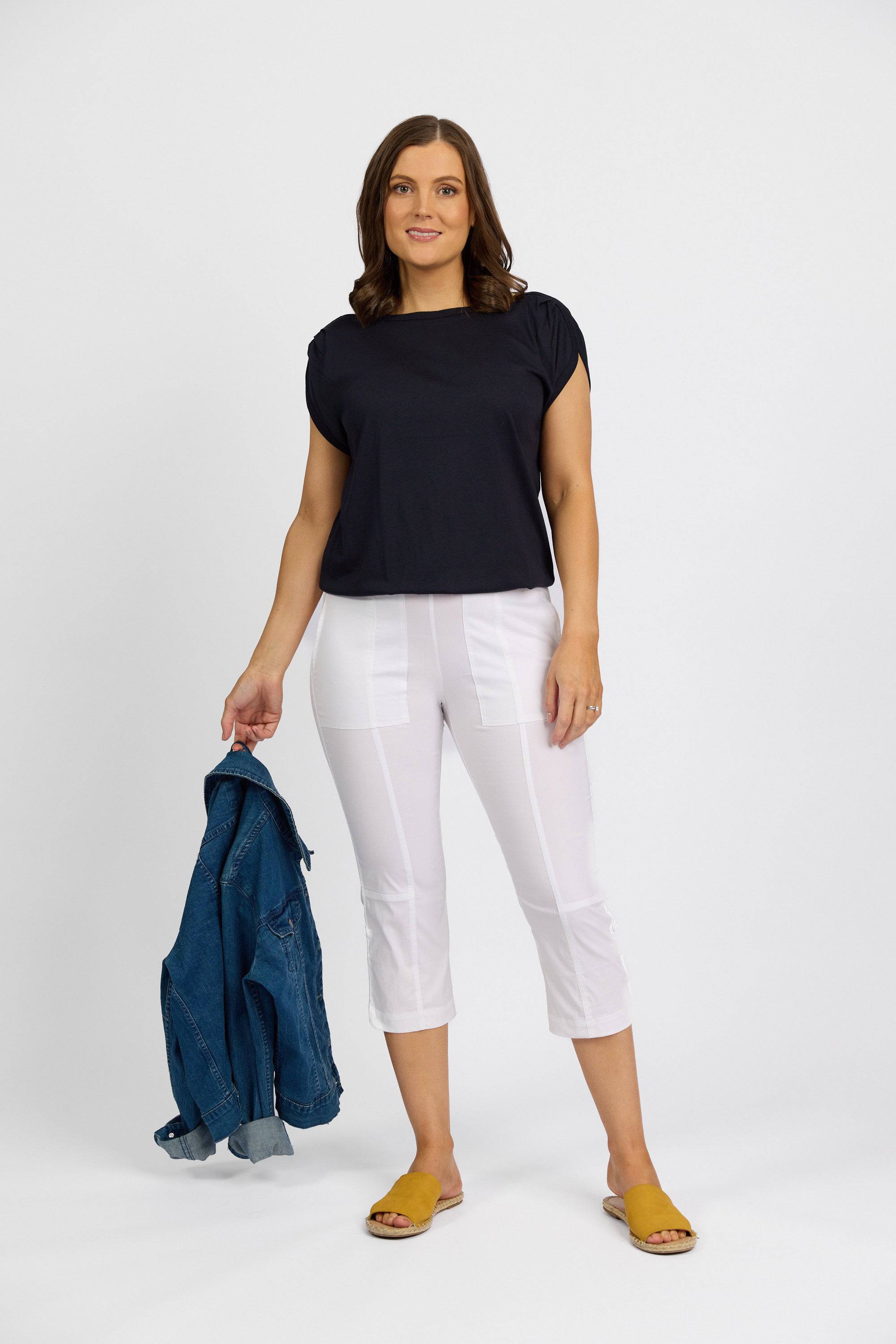 A woman stands against a plain background, wearing a black short-sleeve top, Vassalli Paris 3/4 Cargo Short in white stretch fabric, yellow sandals, and holding a blue denim jacket in her left hand while smiling softly at the camera.