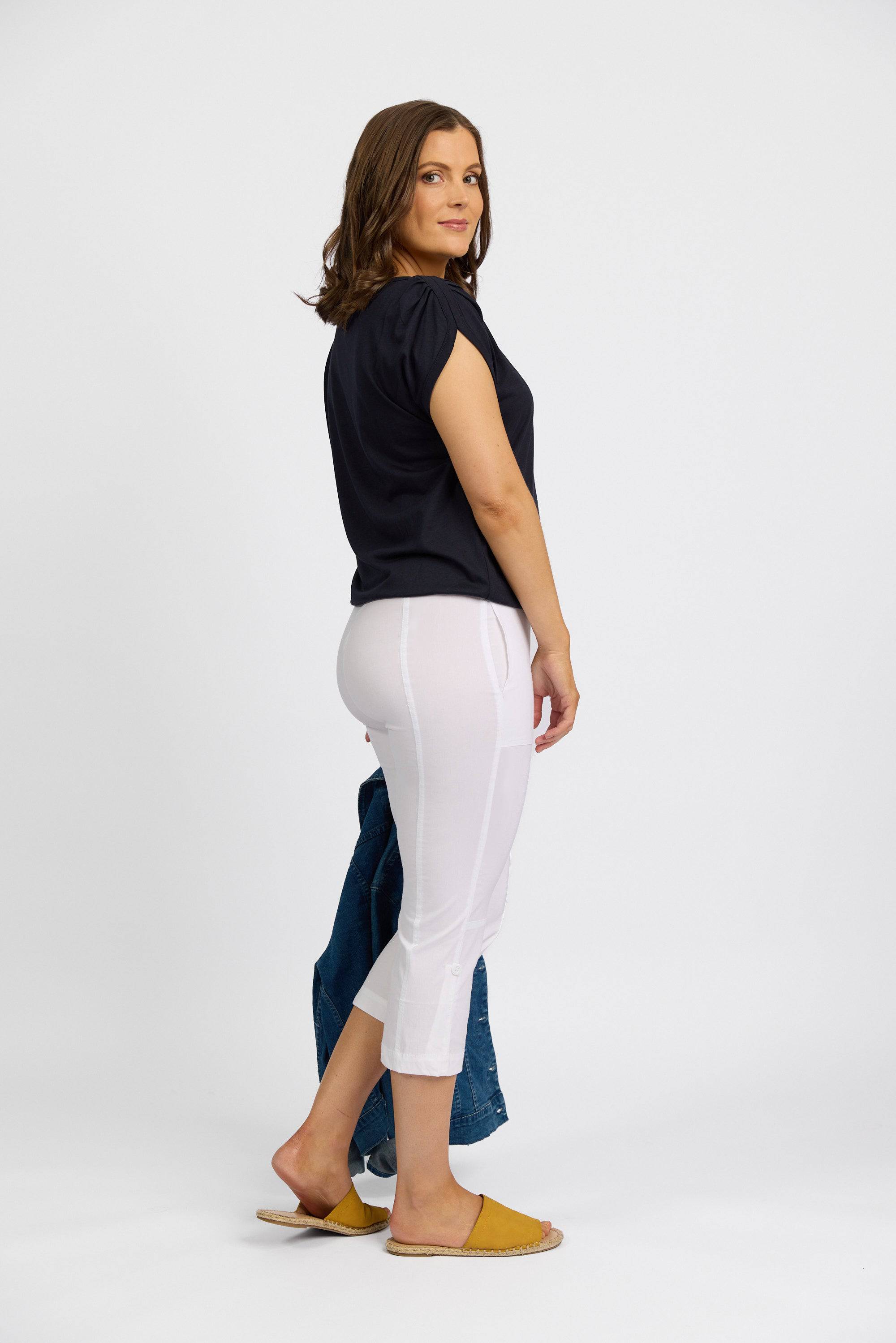 A woman poses sideways in Vassalli's Paris 3/4 Cargo Short - White paired with a black top, yellow slippers, and holds a blue denim jacket, set against a plain white background.