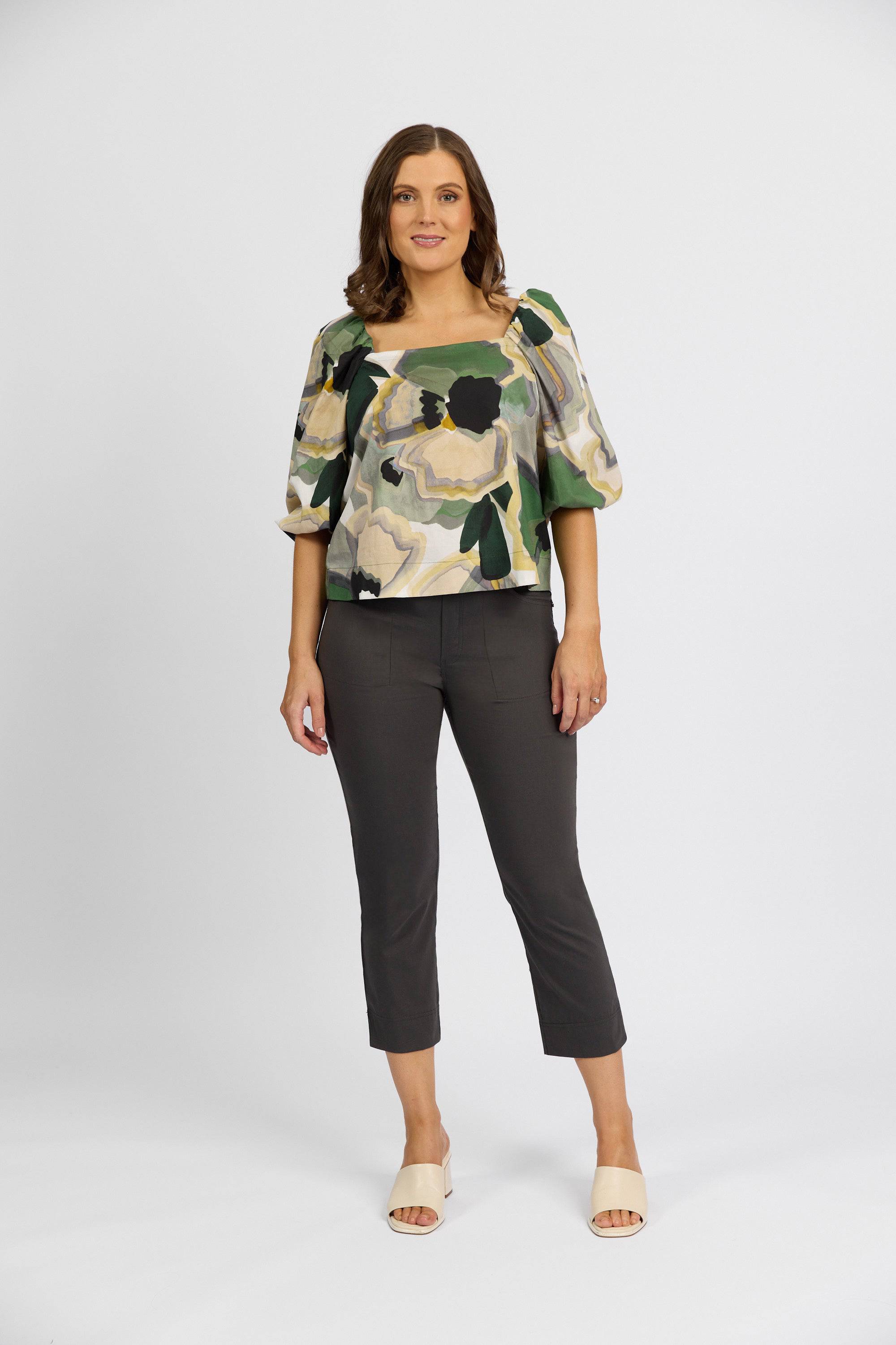 A woman with medium-length brown hair smiles against a white background, wearing a Vassalli green, yellow, and black floral top with puffed sleeves, Paris Tour Capri - Beluga pants by Vassalli, and light-colored sandals.