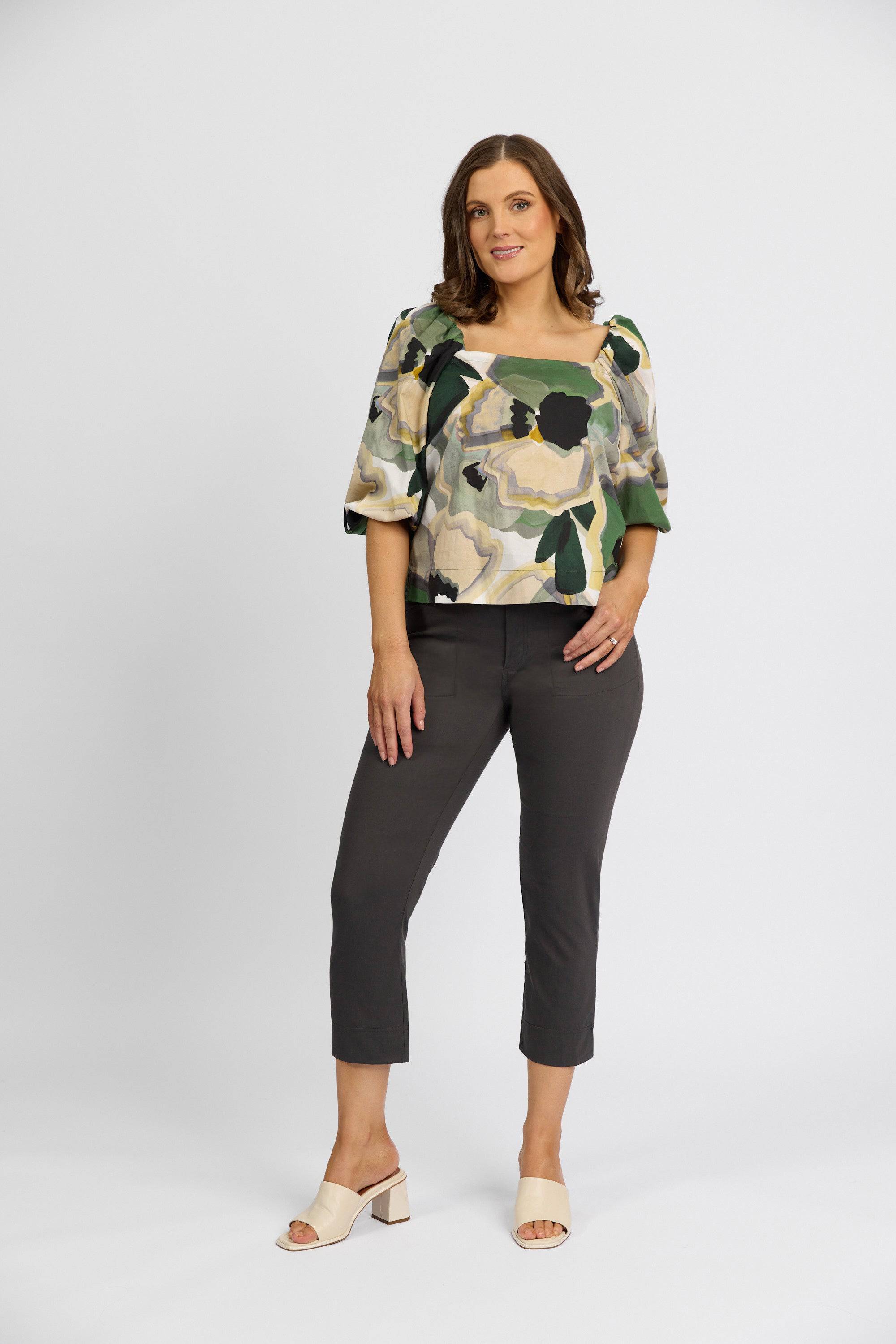 A woman stands against a plain white background wearing a floral print top, Vassalli Paris Tour Capri - Beluga pants, and light-colored heeled sandals. She has one hand on her hip and is smiling slightly.