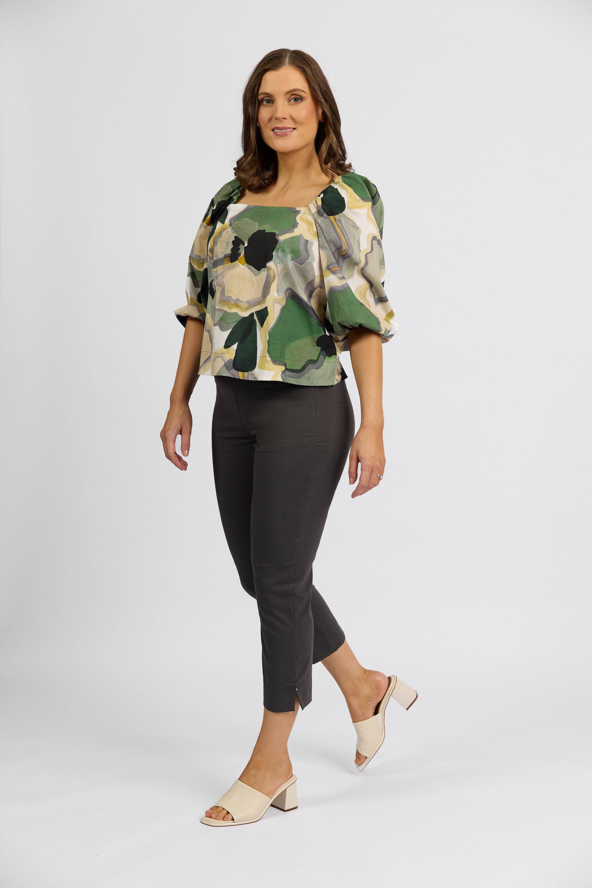 A woman with shoulder-length brown hair smiles against a white background, wearing a Vassalli patterned stretch blouse in green and beige, Paris Tour Capri - Beluga pants by Vassalli, and light heeled sandals.